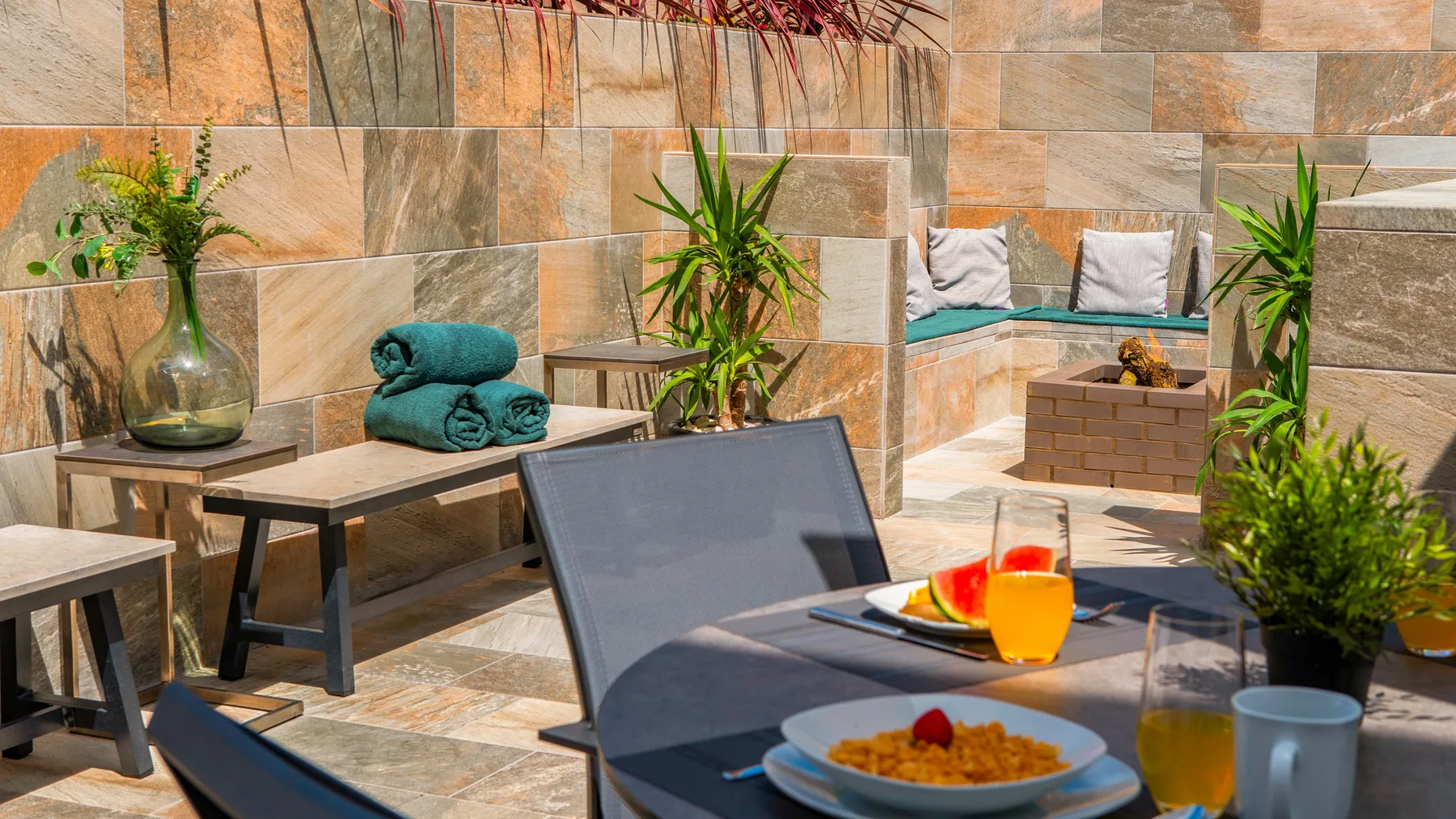 Patio exterior con azulejos, asientos, plantas y una mesa con comida y bebidas.