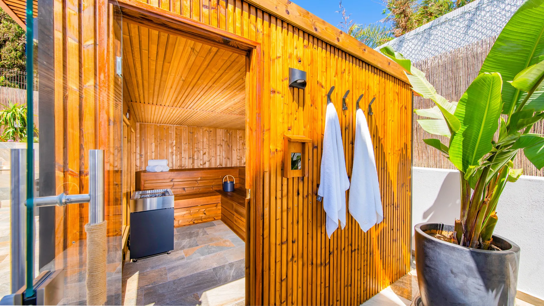 Una sauna exterior de madera da a un patio junto a una planta grande en maceta.