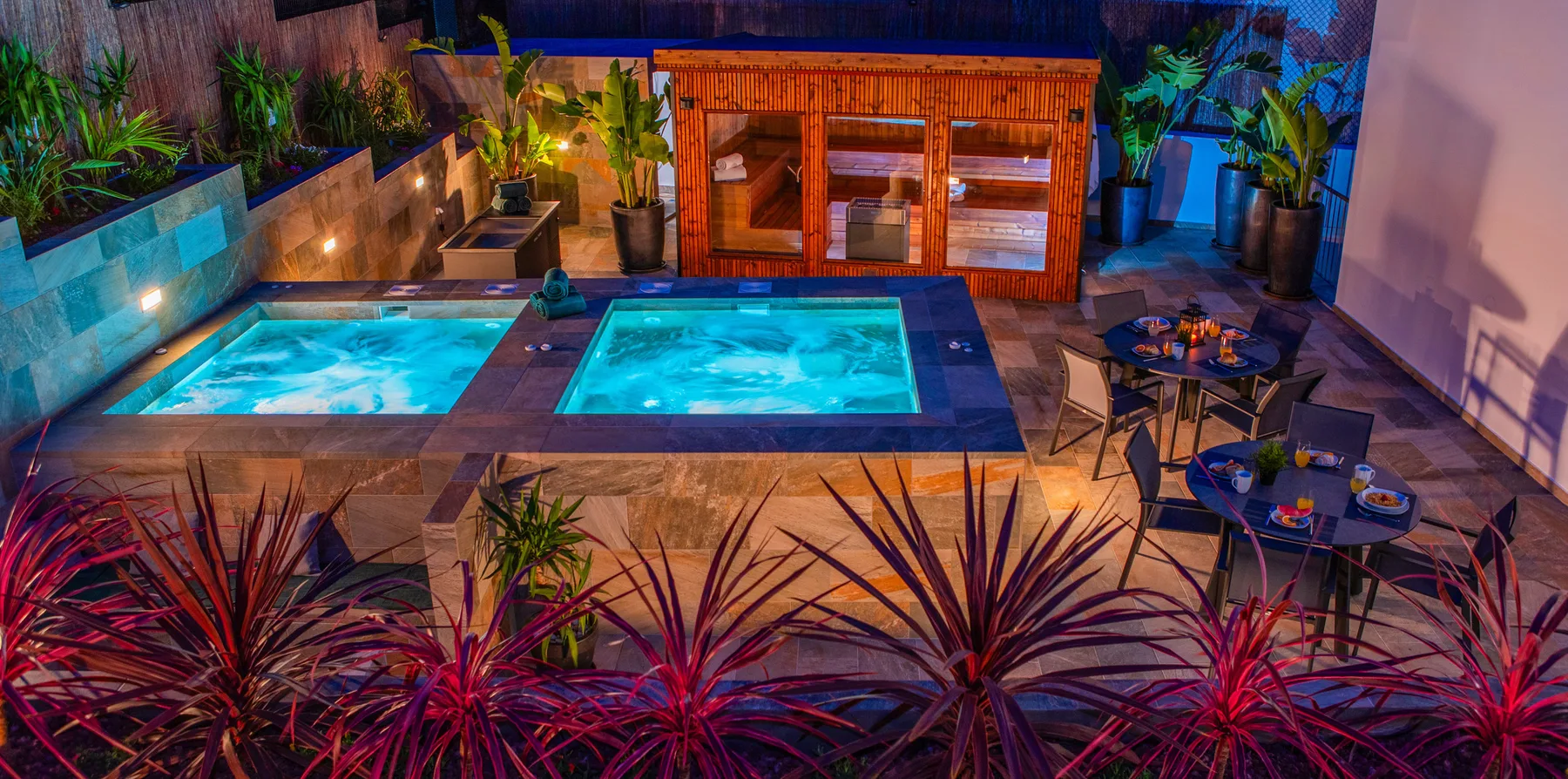 Zona de piscina al aire libre con piscinas iluminadas, asientos y plantas tropicales.