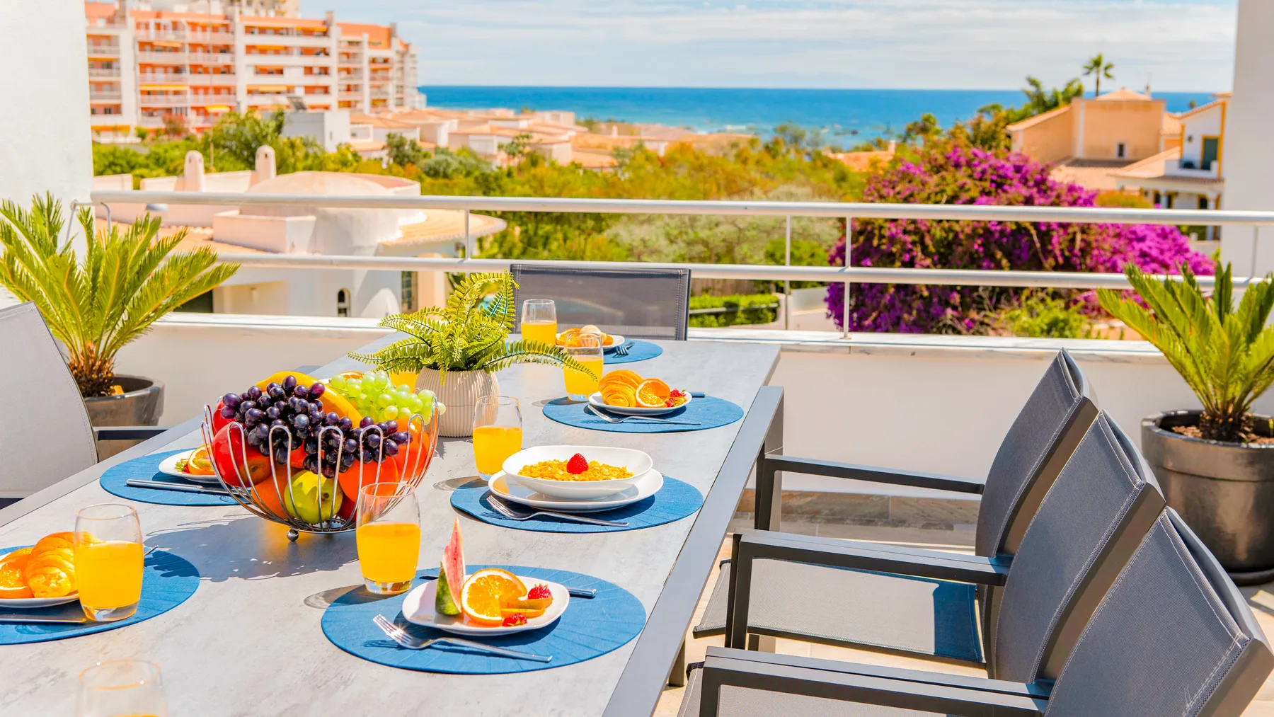 Una mesa de comedor al aire libre está puesta con platos y fruta, con vistas al mar.