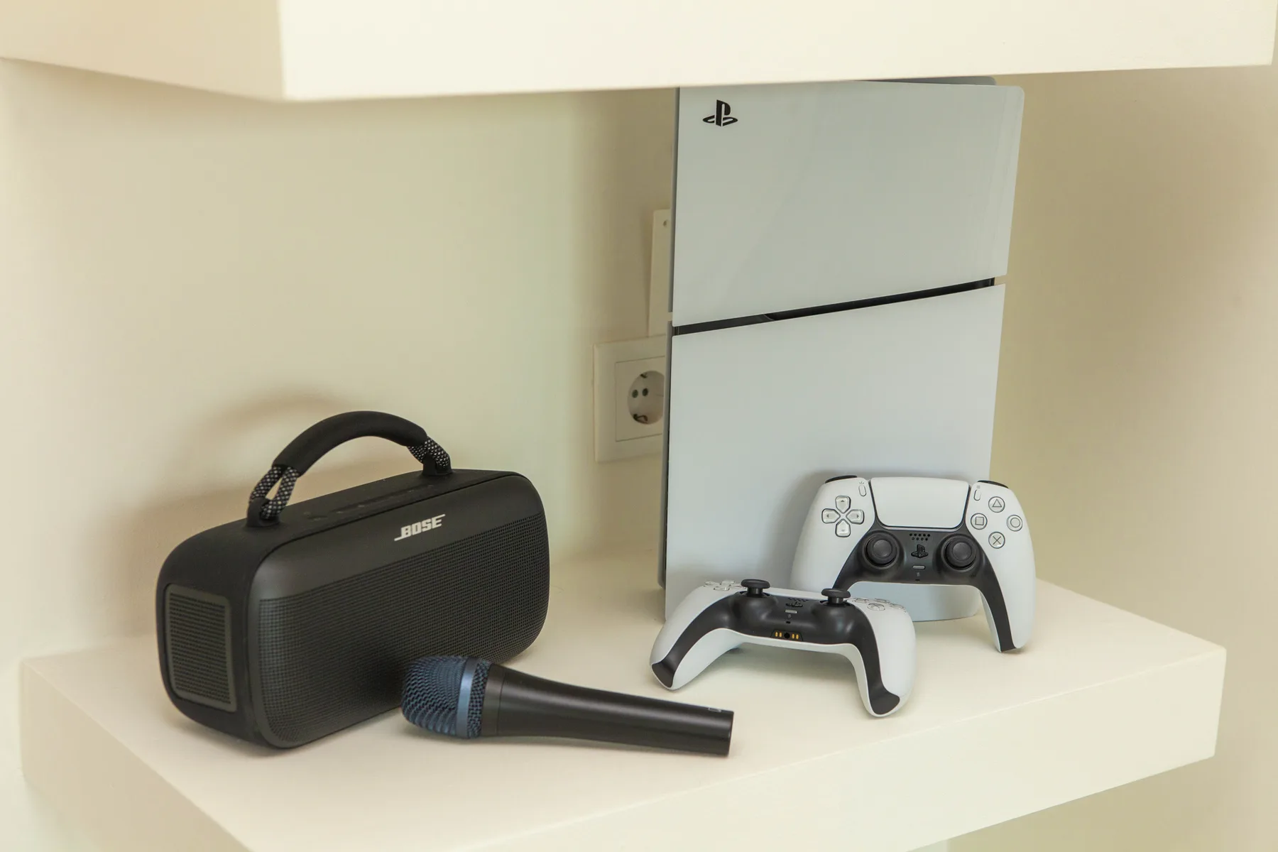 Una consola de videojuegos, mandos y un altavoz están sobre un estante blanco.