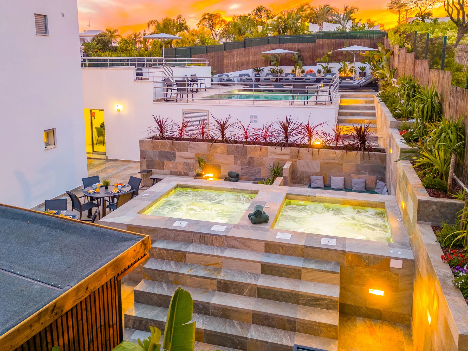 Una piscina exterior y jacuzzis iluminados están rodeados de asientos y plantas tropicales.