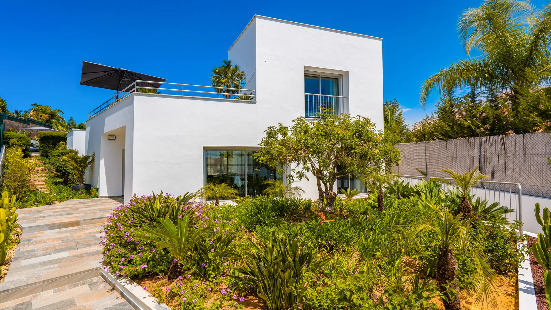 Una casa blanca moderna se alza junto a un jardín paisajístico bajo un cielo azul despejado.