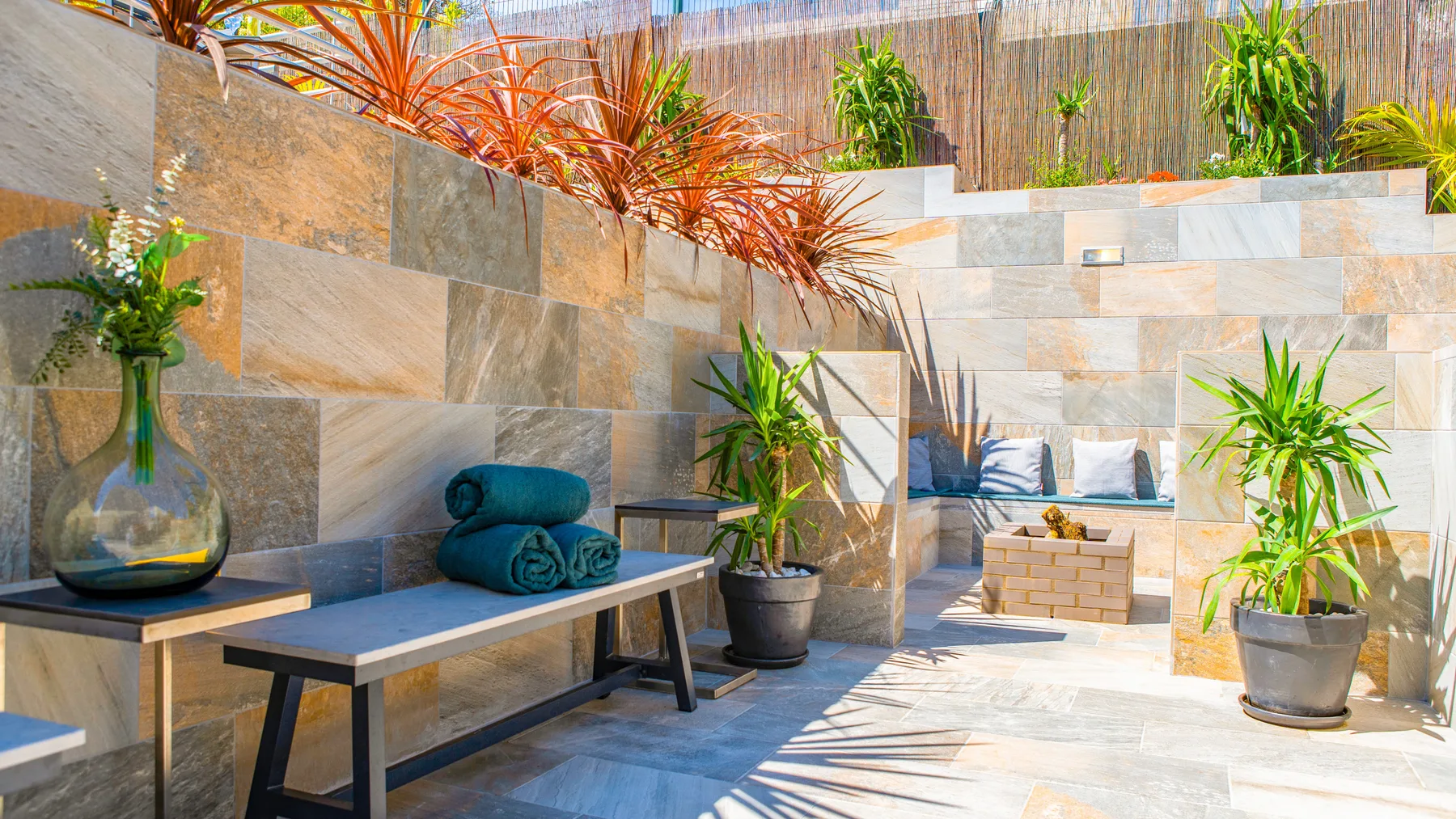 Un patio soleado con azulejos, un banco, plantas en macetas y un rincón con asientos acolchados.