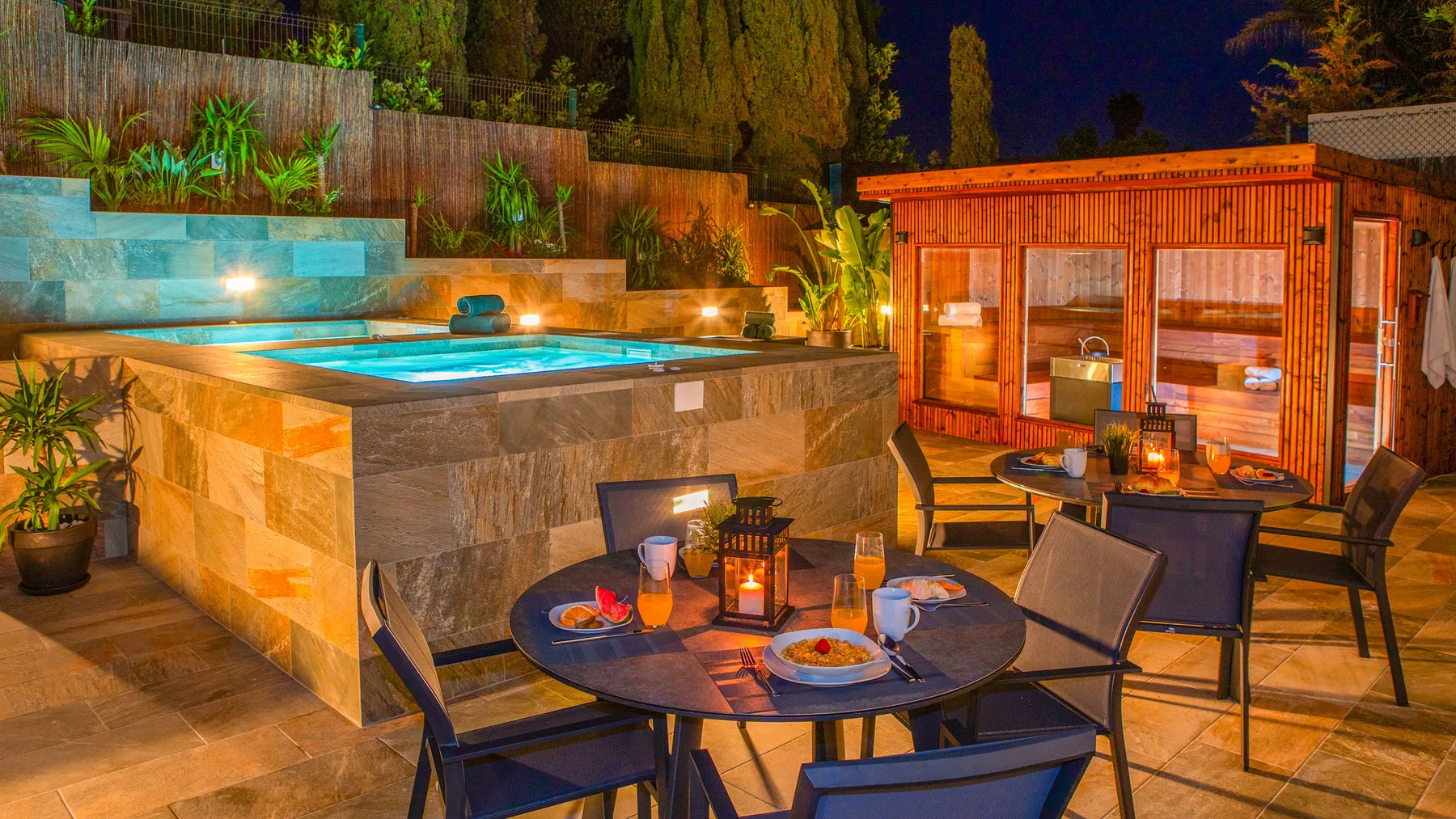 Un patio nocturno cuenta con una bañera de hidromasaje iluminada, asientos al aire libre y una cabaña de madera.