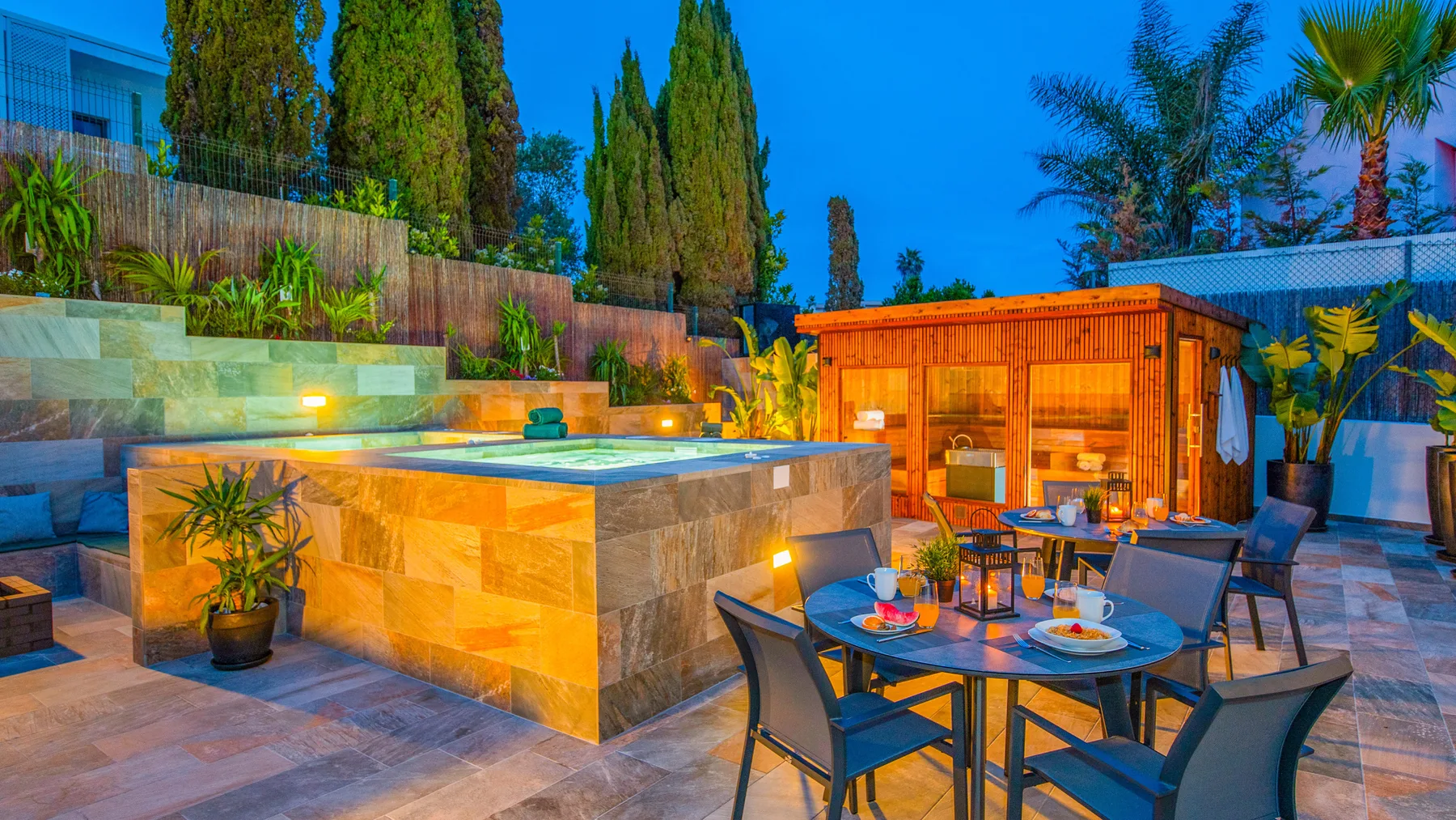 Un patio exterior cuenta con una bañera de hidromasaje elevada, una mesa de comedor y una sauna de madera.