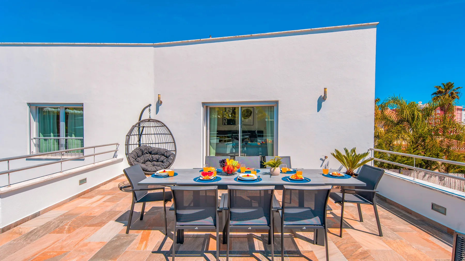 Una zona de comedor en la terraza tiene una mesa grande y sillas junto a un edificio blanco.