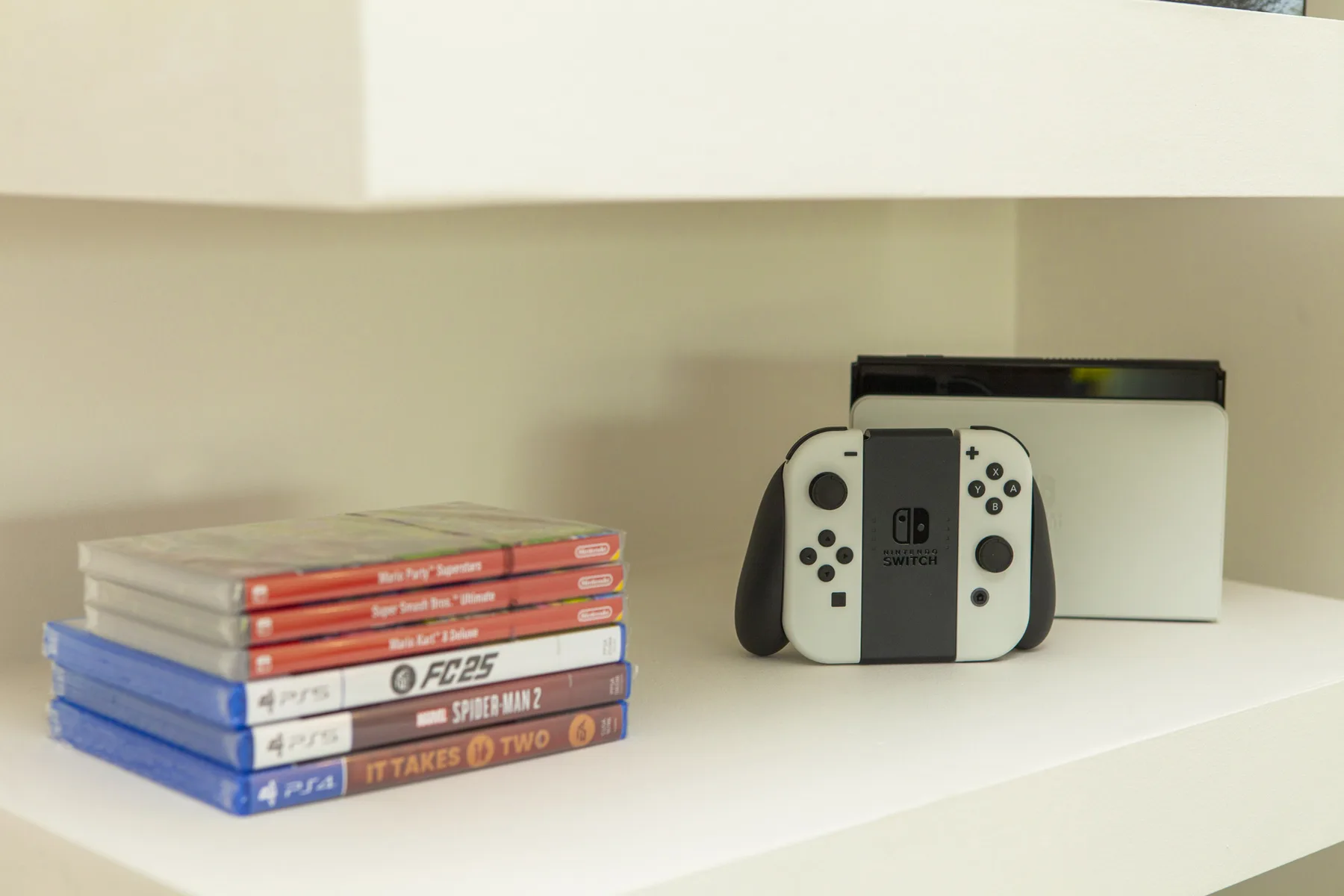 Una consola de videojuegos y mandos están junto a una pila de cajas de juegos en un estante.