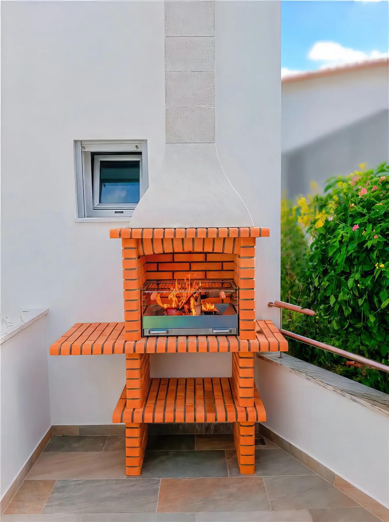 Una barbacoa de ladrillo al aire libre está junto a una pared blanca y vegetación.