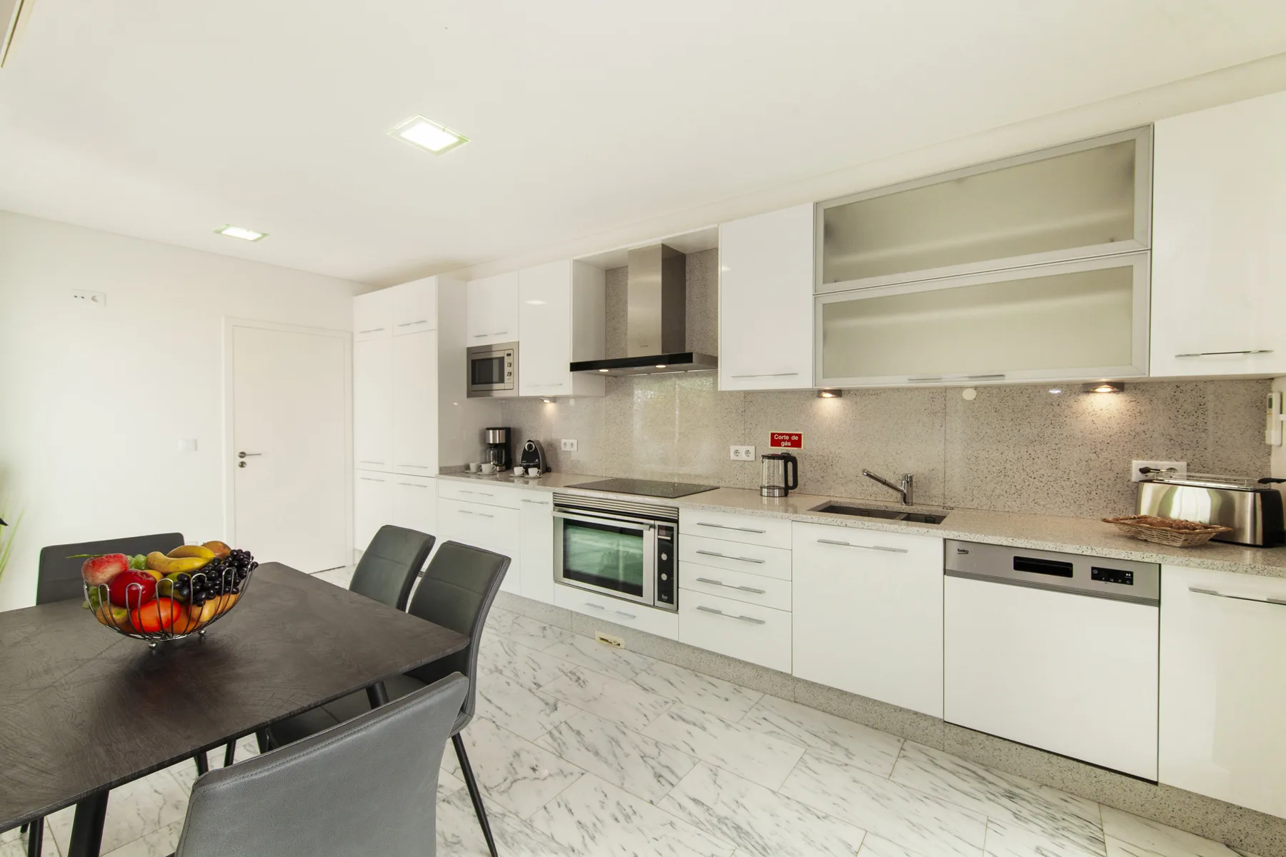 Una cocina luminosa con comedor, gabinetes blancos y una mesa oscura.