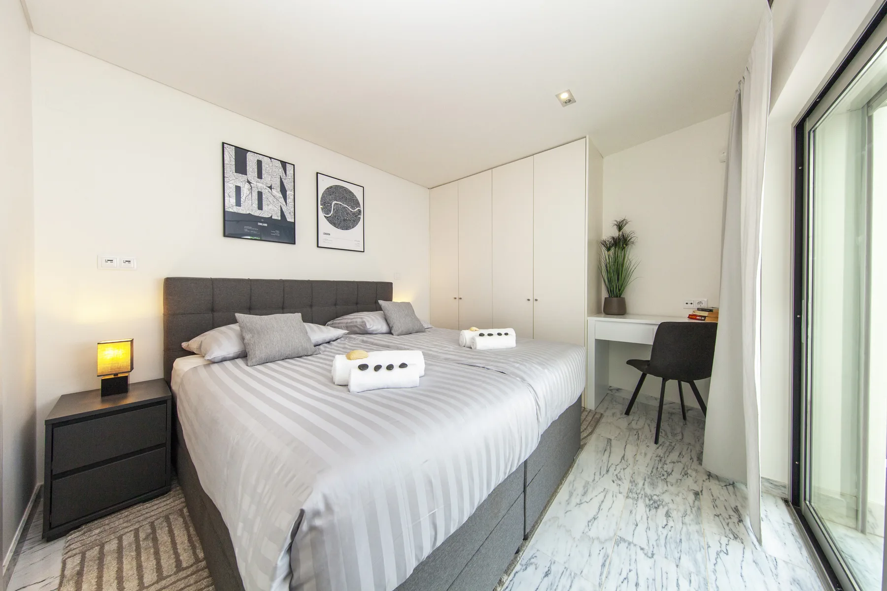 Un dormitorio luminoso con cama doble, mesita de noche y escritorio junto a la ventana.