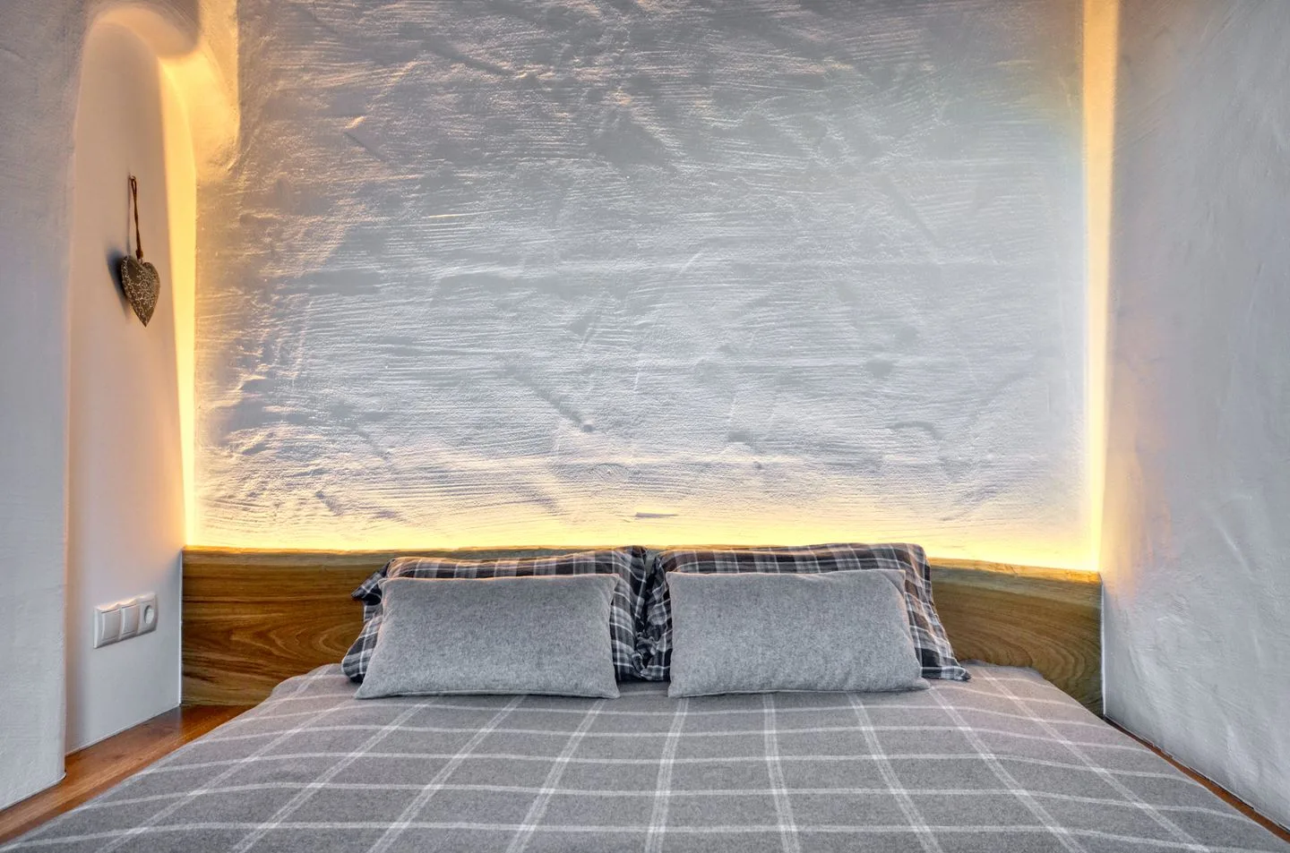 Una cama con ropa de cama gris está frente a una pared texturizada con iluminación suave.