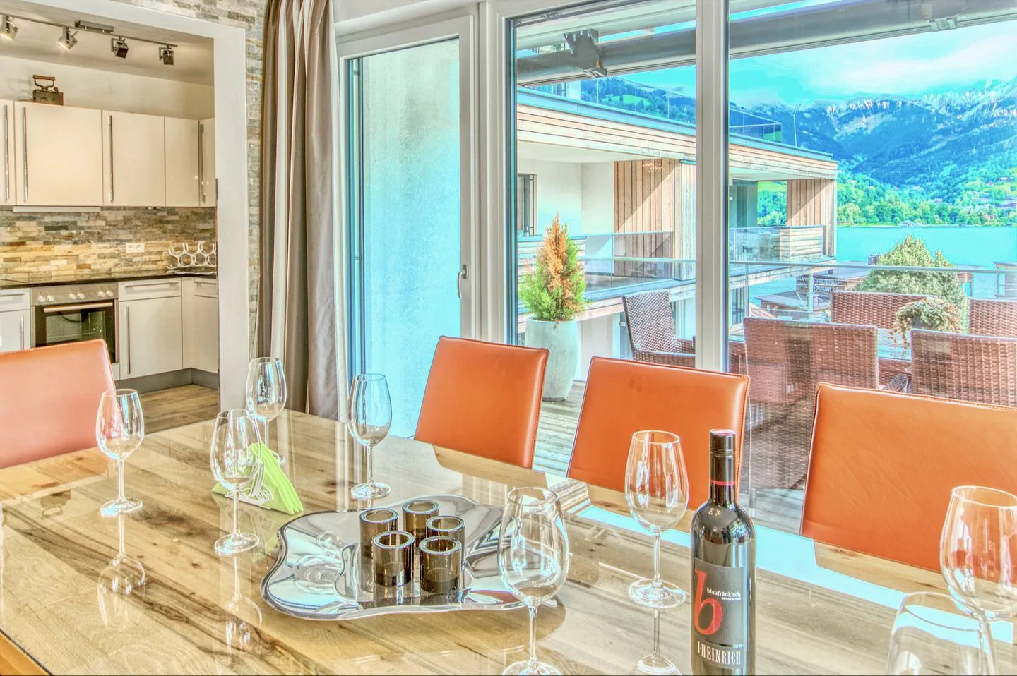 Una mesa de comedor con asientos naranjas está junto a grandes ventanales con vistas a un lago y montañas.