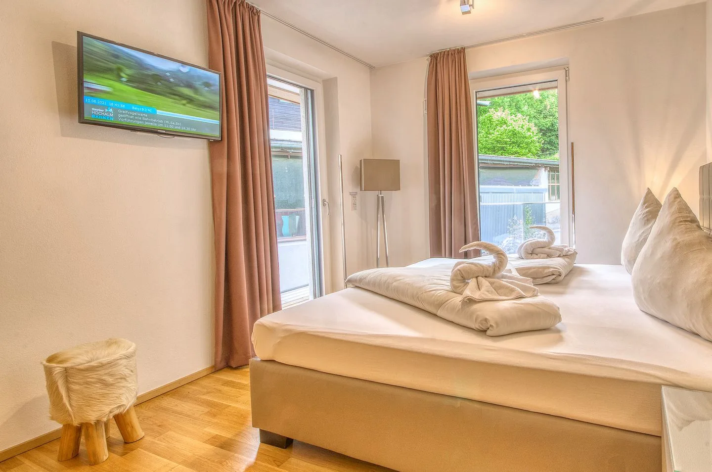 Un dormitorio luminoso con una cama grande, ventanas con cortinas y un televisor en la pared.