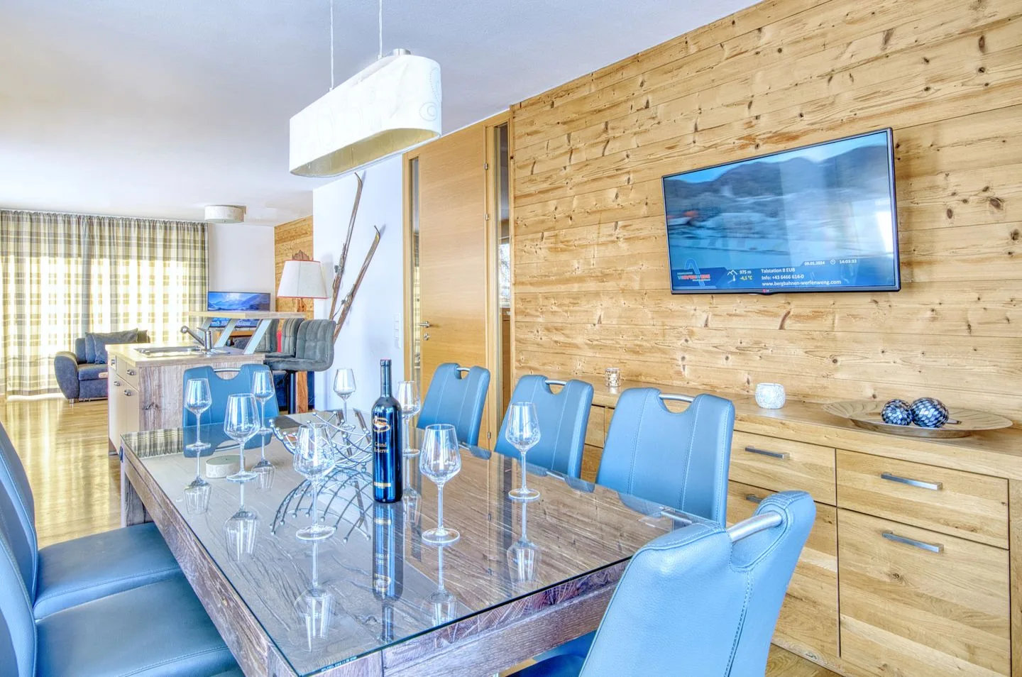 Un comedor con asientos azules, una mesa con cubierta de vidrio y un televisor en la pared revestida de madera.
