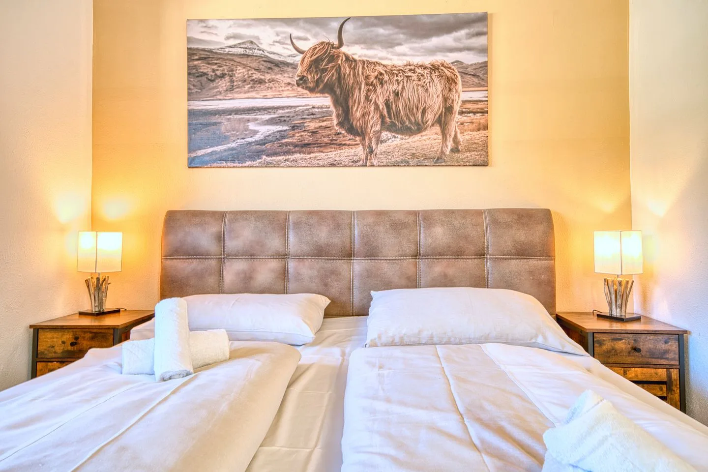 A bedroom with a bed beneath a large painting of a shaggy cow.