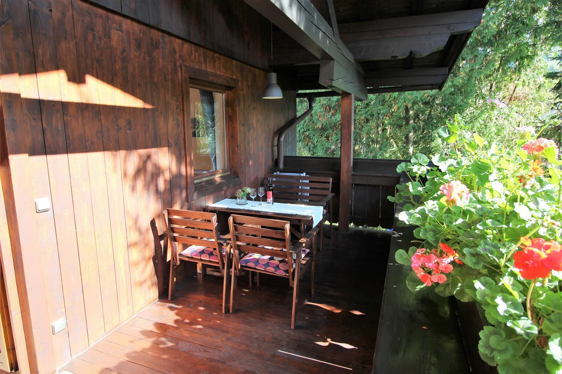 A covered wooden patio has a dining table and seating beside flowering plants.