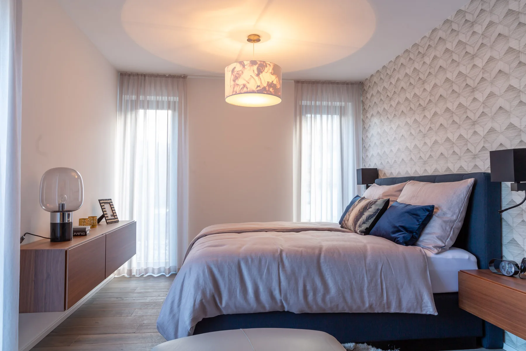 A bedroom with a neatly made bed and tall windows with sheer curtains.