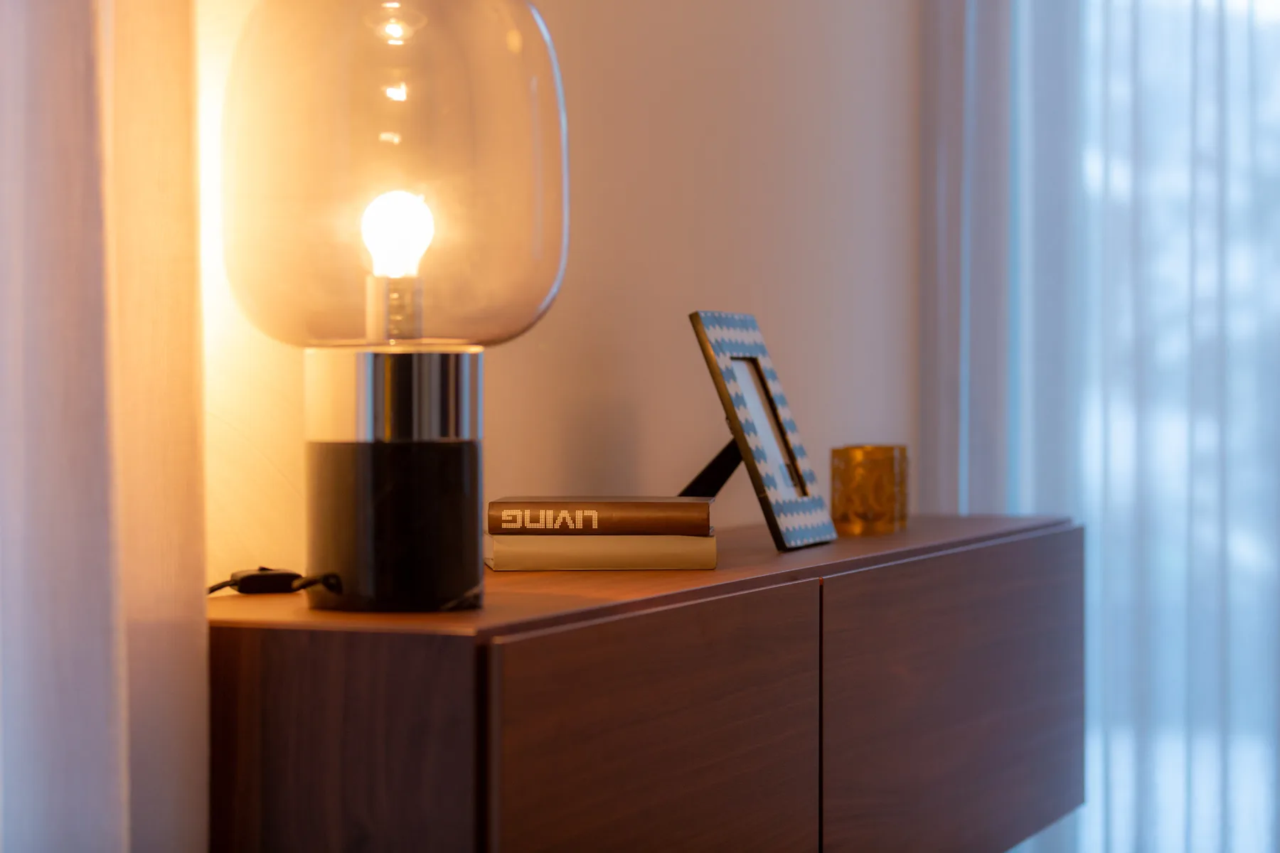 A wooden cabinet holds a glowing table lamp beside a window.