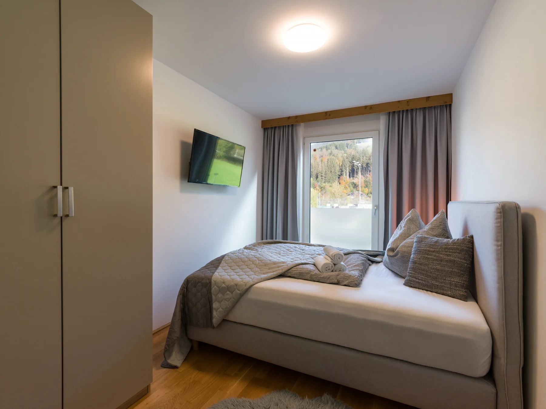 A bedroom with a neatly made bed, bedside window, and wall-mounted television.