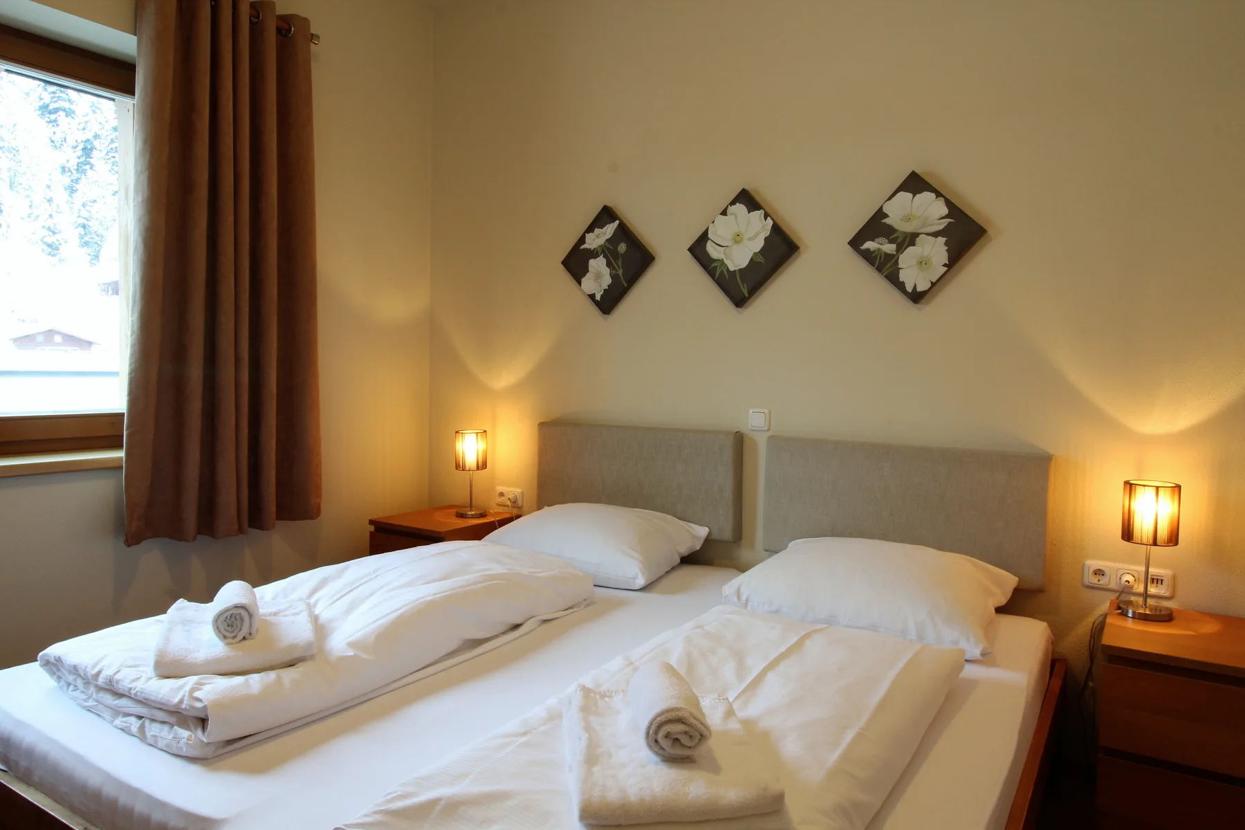 A bedroom with two beds, folded towels, bedside lamps, and floral wall art.