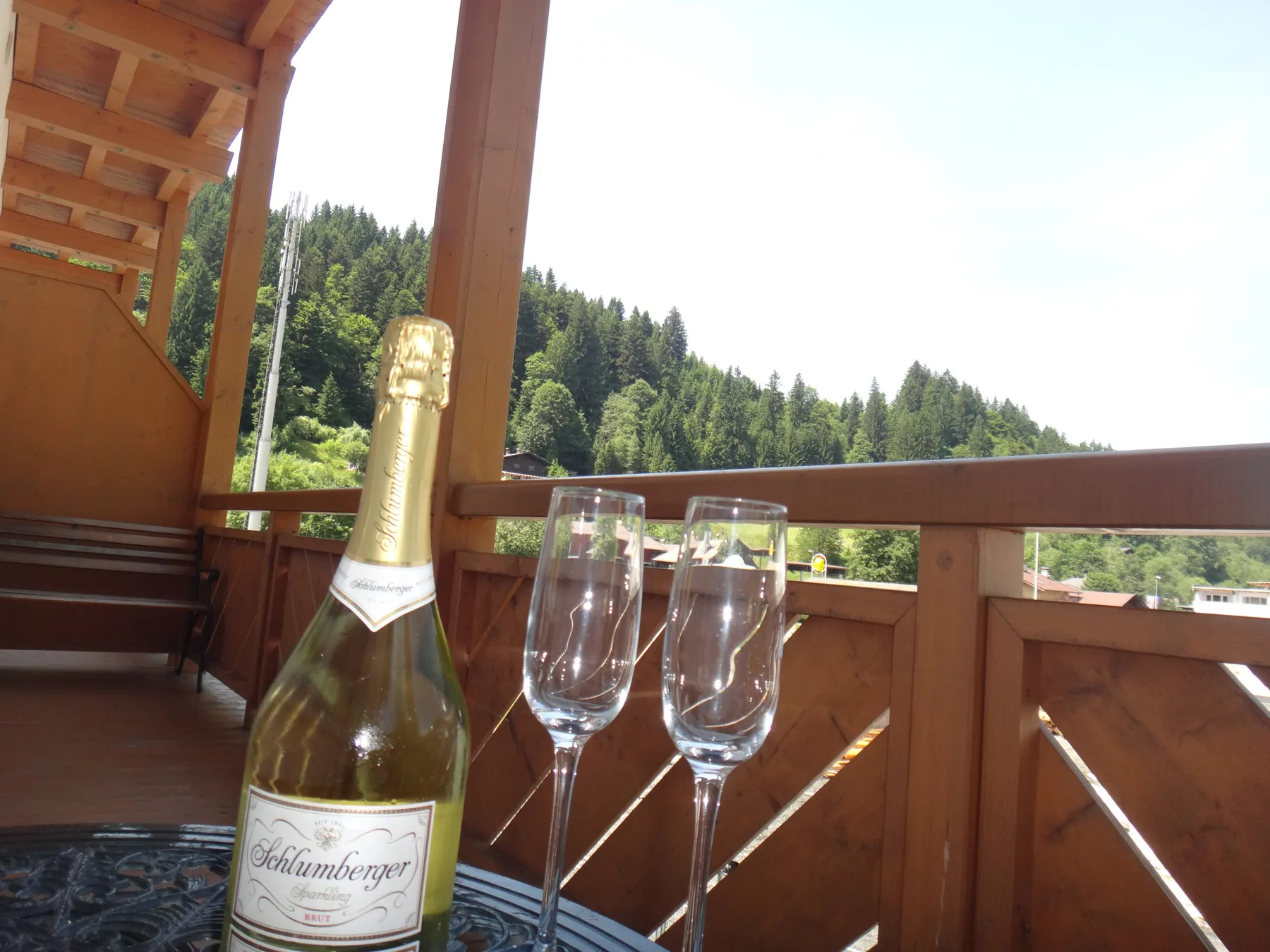 A wooden balcony overlooks a forested hillside, with a bottle and glasses on the railing.