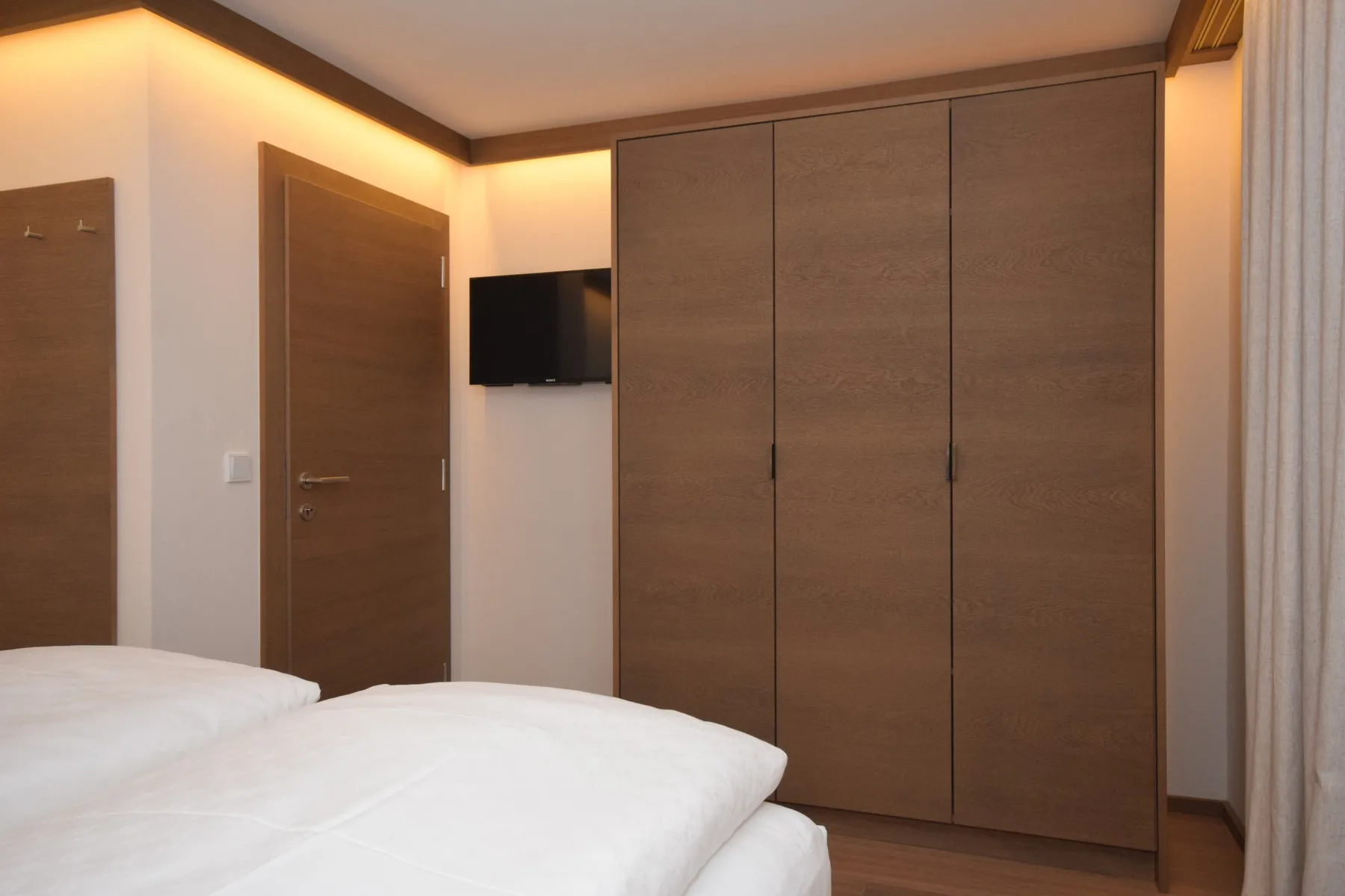 Dormitorio con ropa de cama blanca, un armario y un televisor en la pared.