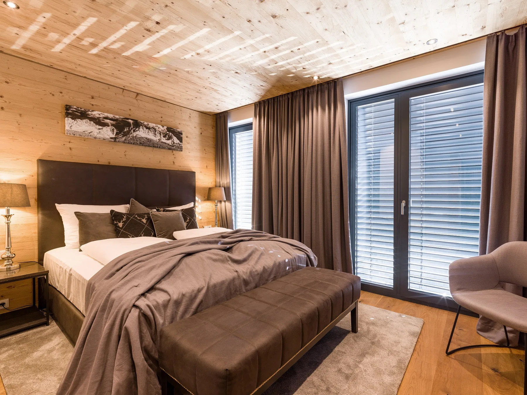 Un dormitorio con una cama grande, un banco y ventanas altas con persianas.
