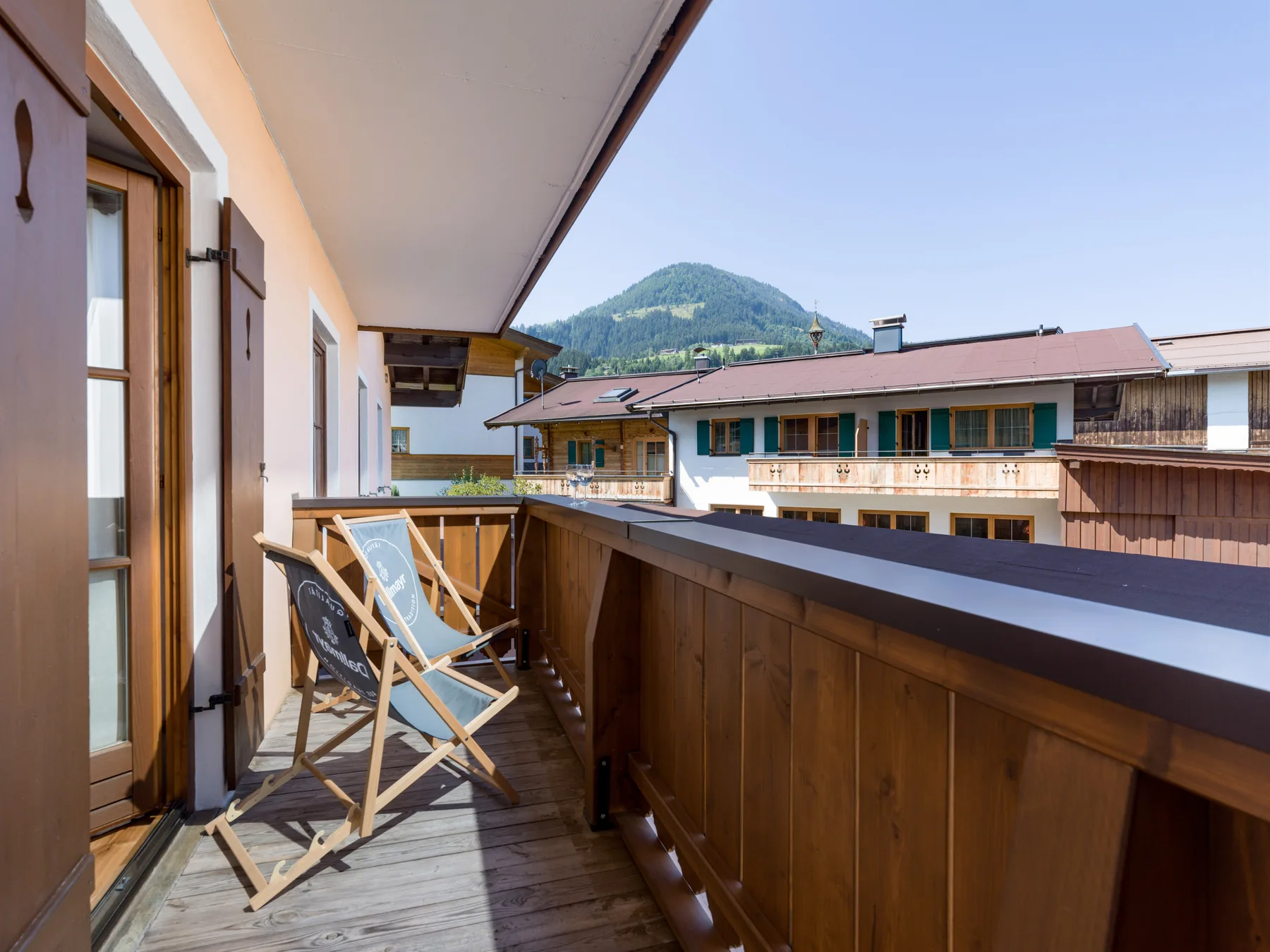 A covered balcony with seating overlooks nearby buildings and a green mountain.