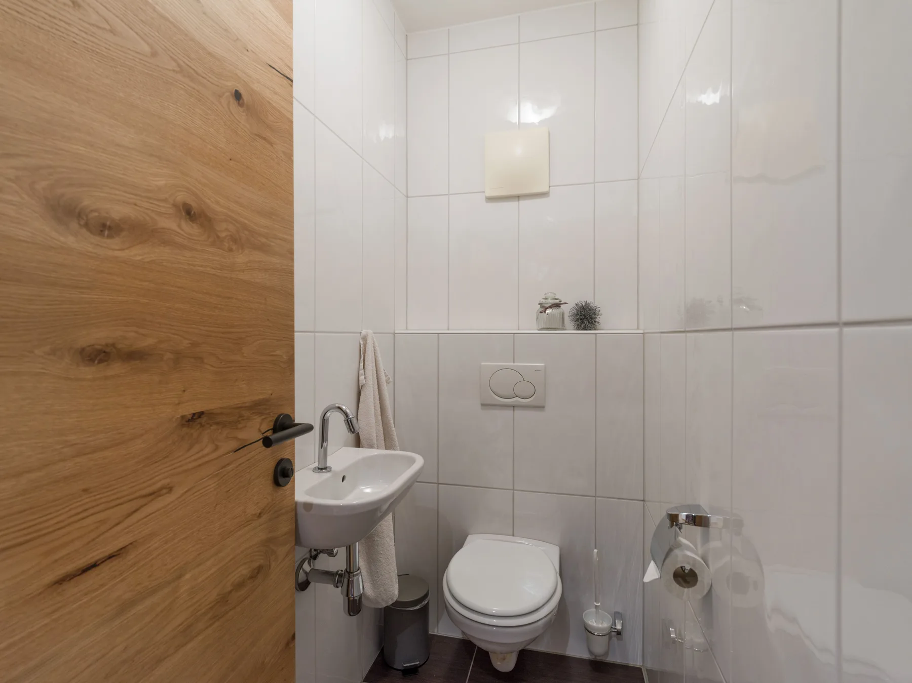 A tiled bathroom with a wall-mounted toilet and a small sink beside a wooden door.