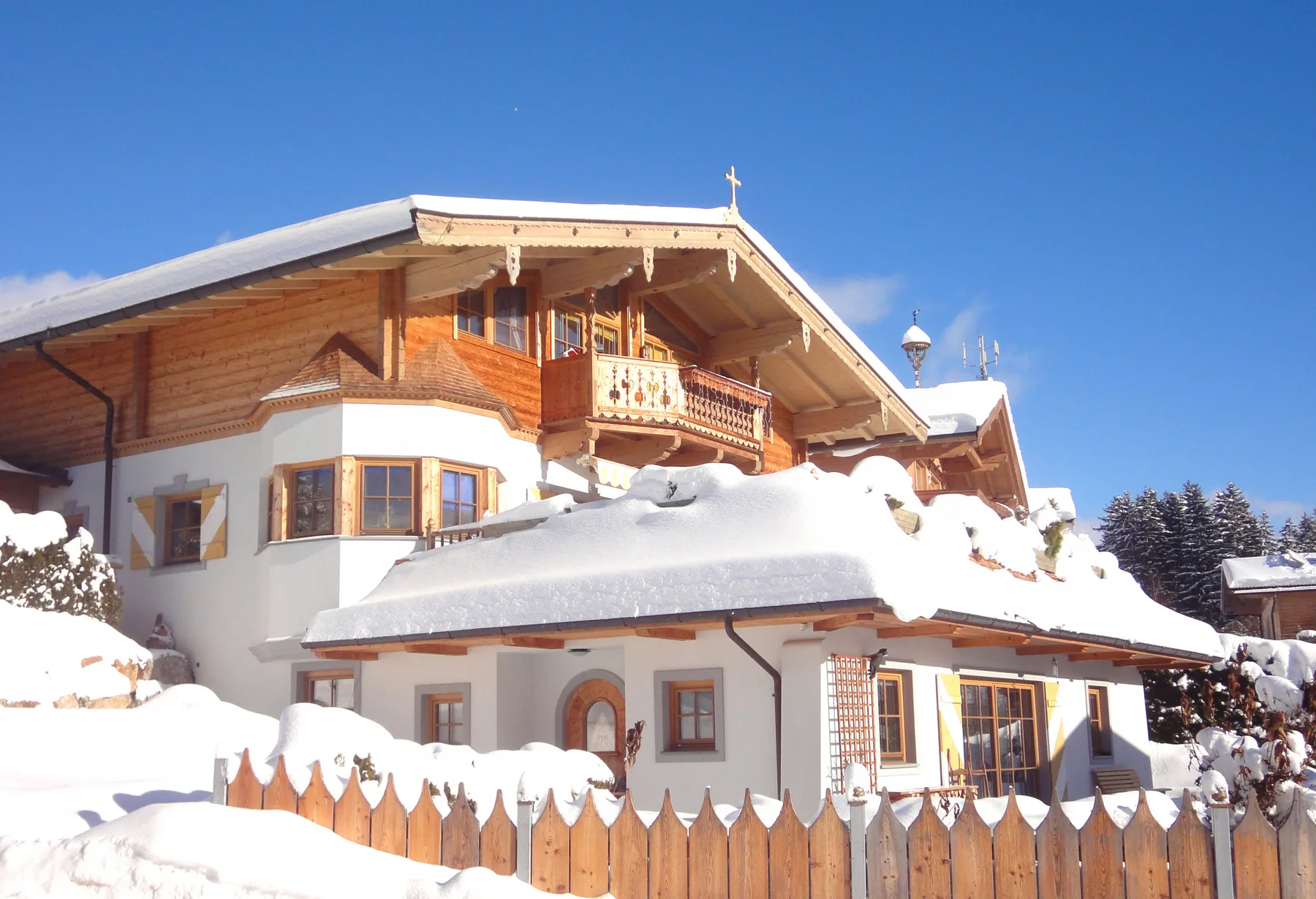 Zona común: Un chalet cubierto de nieve se alza tras una cerca de madera bajo un cielo azul despejado.