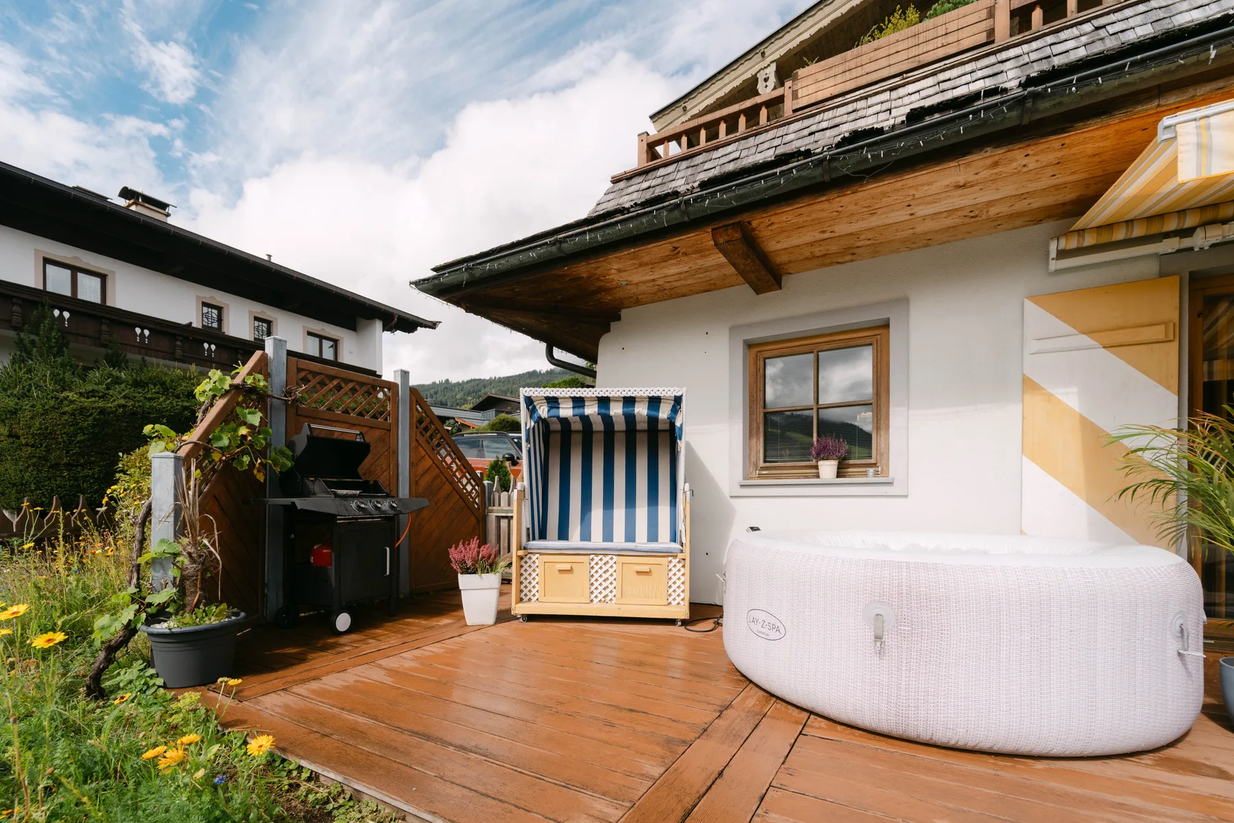 Una terraza exterior de madera con jacuzzi cubierto, biombo a rayas y parrilla.