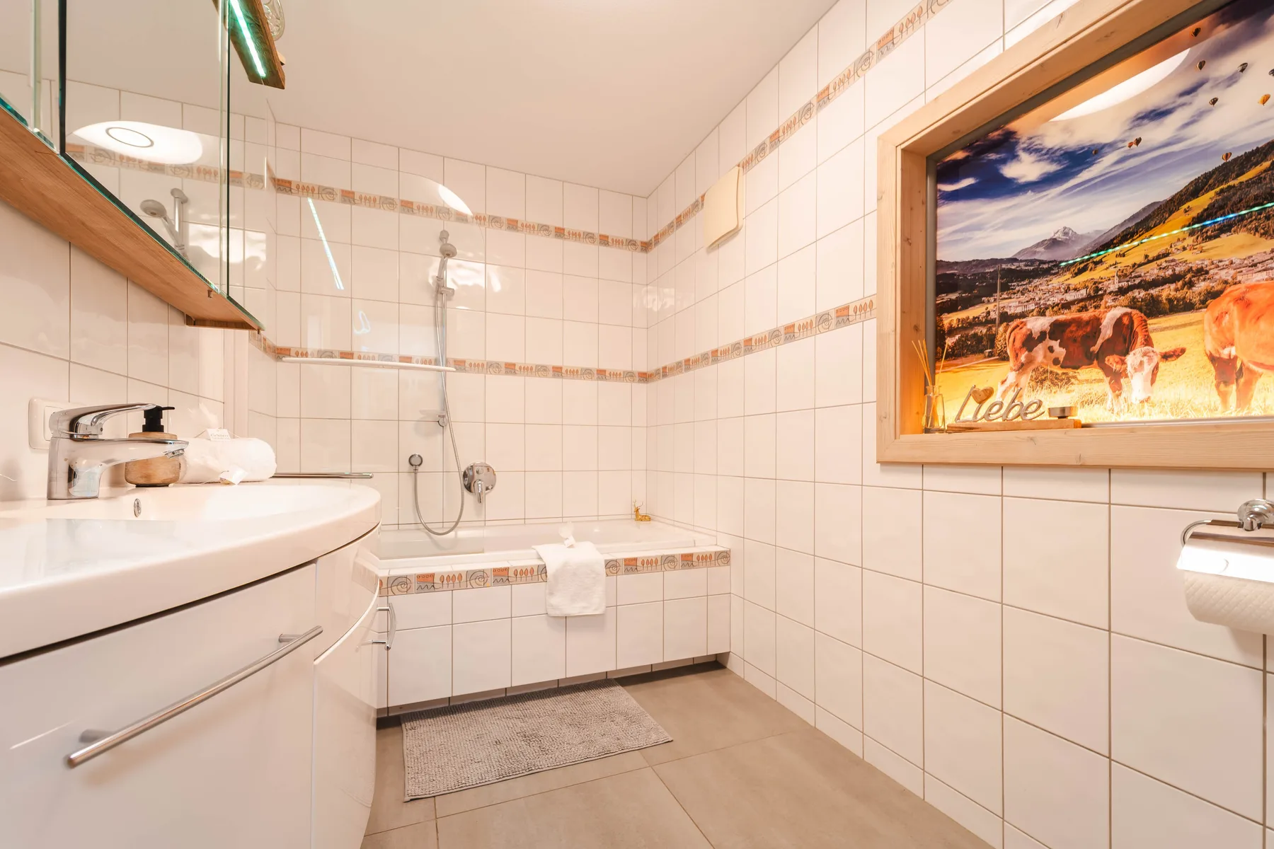 Un baño alicatado con bañera, lavabo y un cuadro de paisaje.