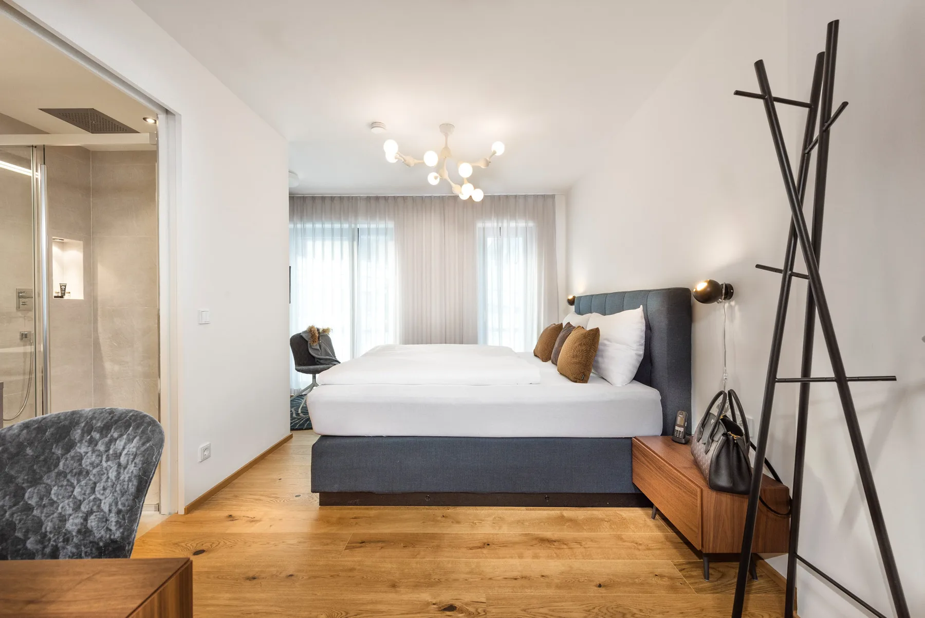 Un dormitorio luminoso con una cama bien tendida, ventanas grandes y suelo de madera.