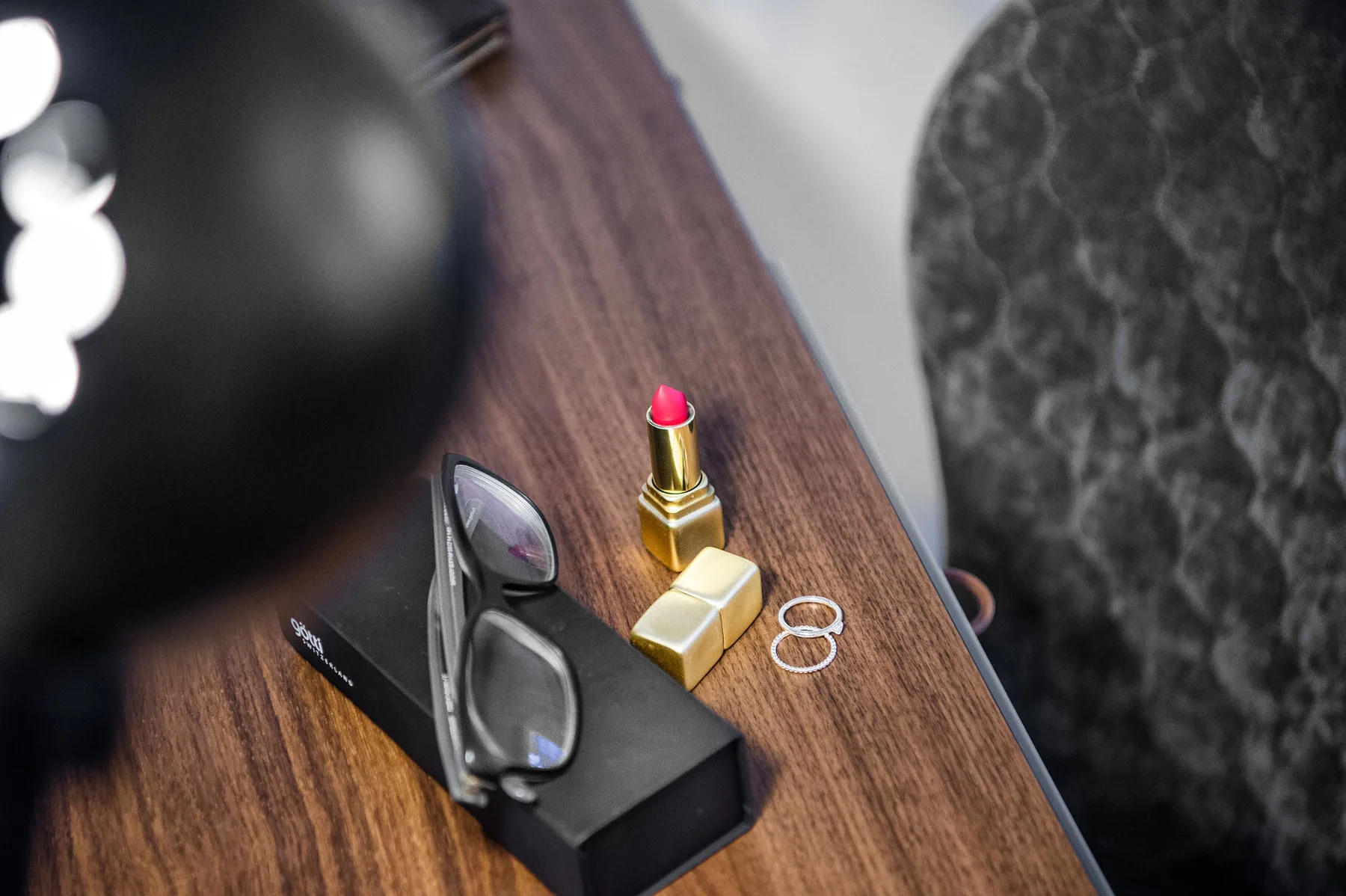 Una mesa de madera sostiene unas gafas, un lápiz labial, una cajita y dos anillos.