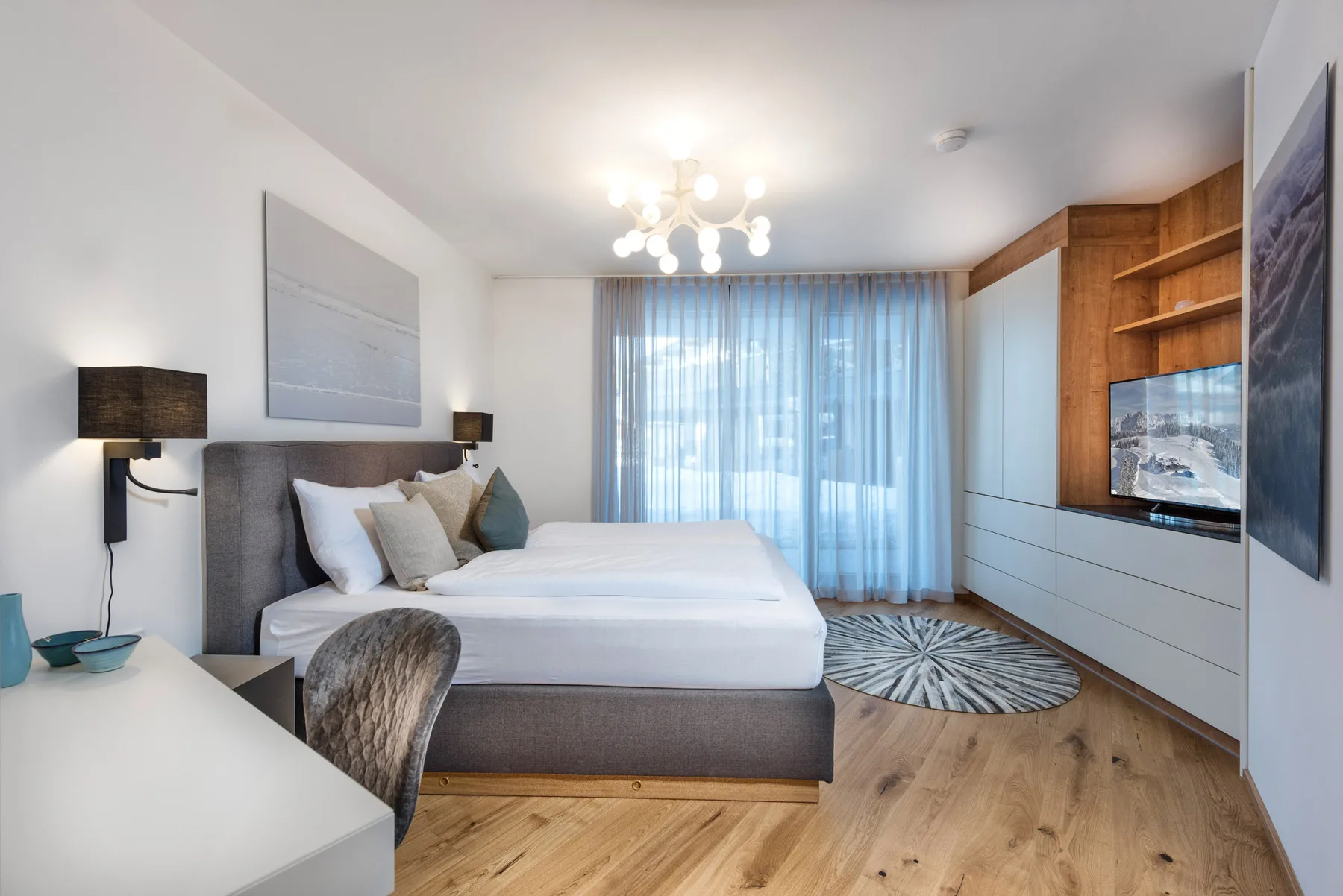Un dormitorio luminoso con una cama grande, cortinas translúcidas y estantes de madera empotrados.