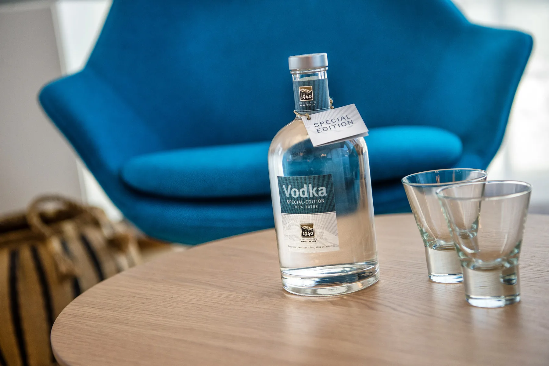 Una botella de vodka y dos vasos están sobre una mesa frente a una silla azul.