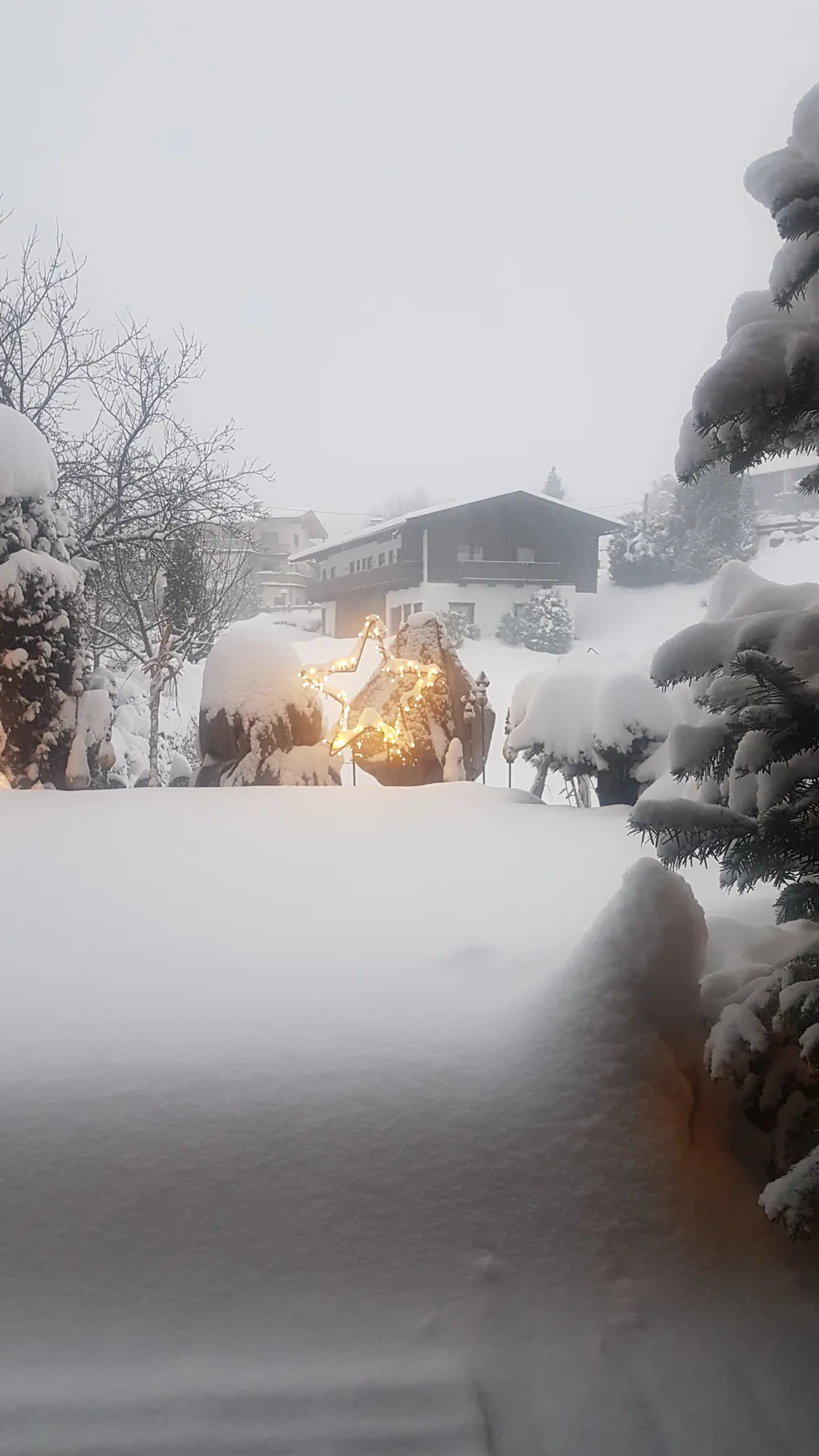 Escena exterior cubierta de nieve, con una casa y un pequeño fuego que brilla entre los árboles.