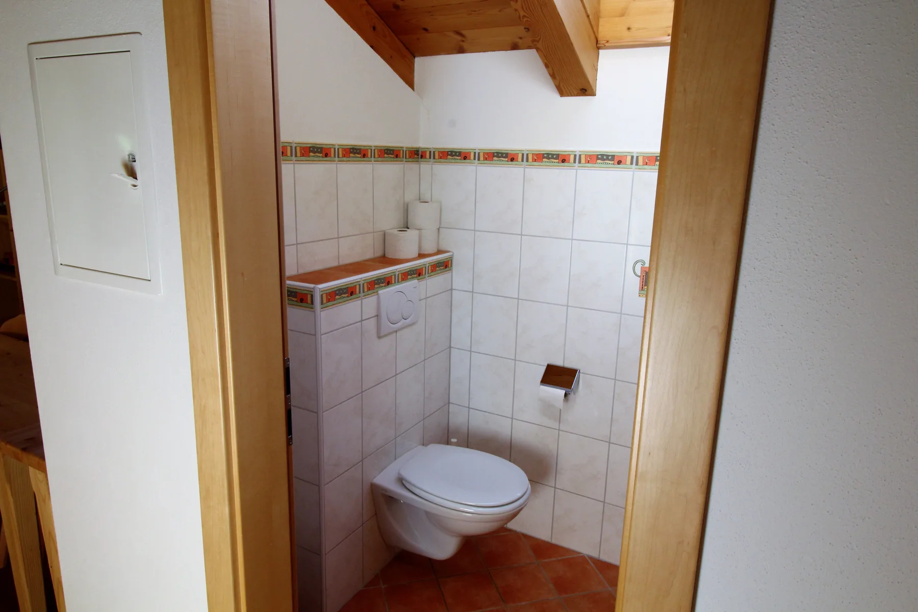 A compact tiled bathroom with a wall-mounted toilet beneath a sloped wooden ceiling.