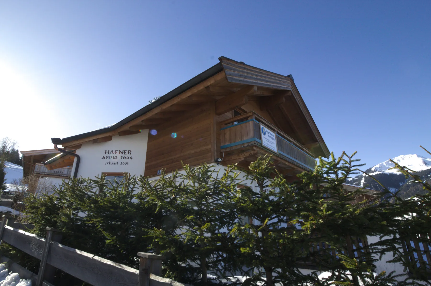 A wooden chalet stands behind snow-dusted shrubs, with mountains in the background.