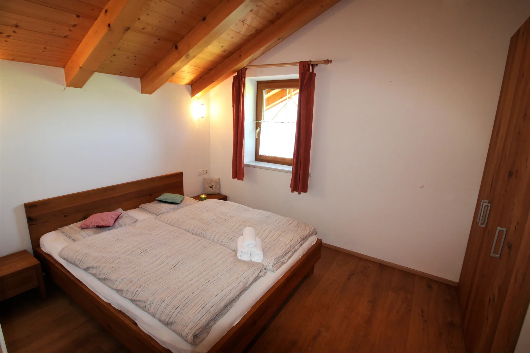 A bedroom with a wooden bed beneath a sloped wood ceiling and a curtained window.