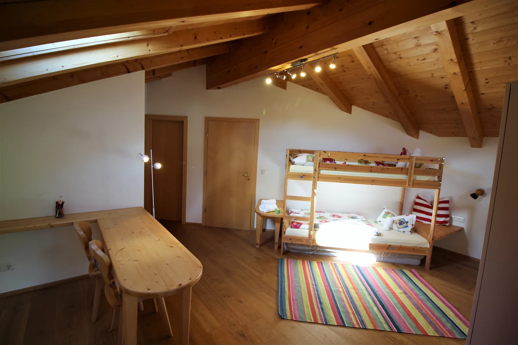 A wood-beamed bedroom with bunk beds, a dining table, and a colorful rug.