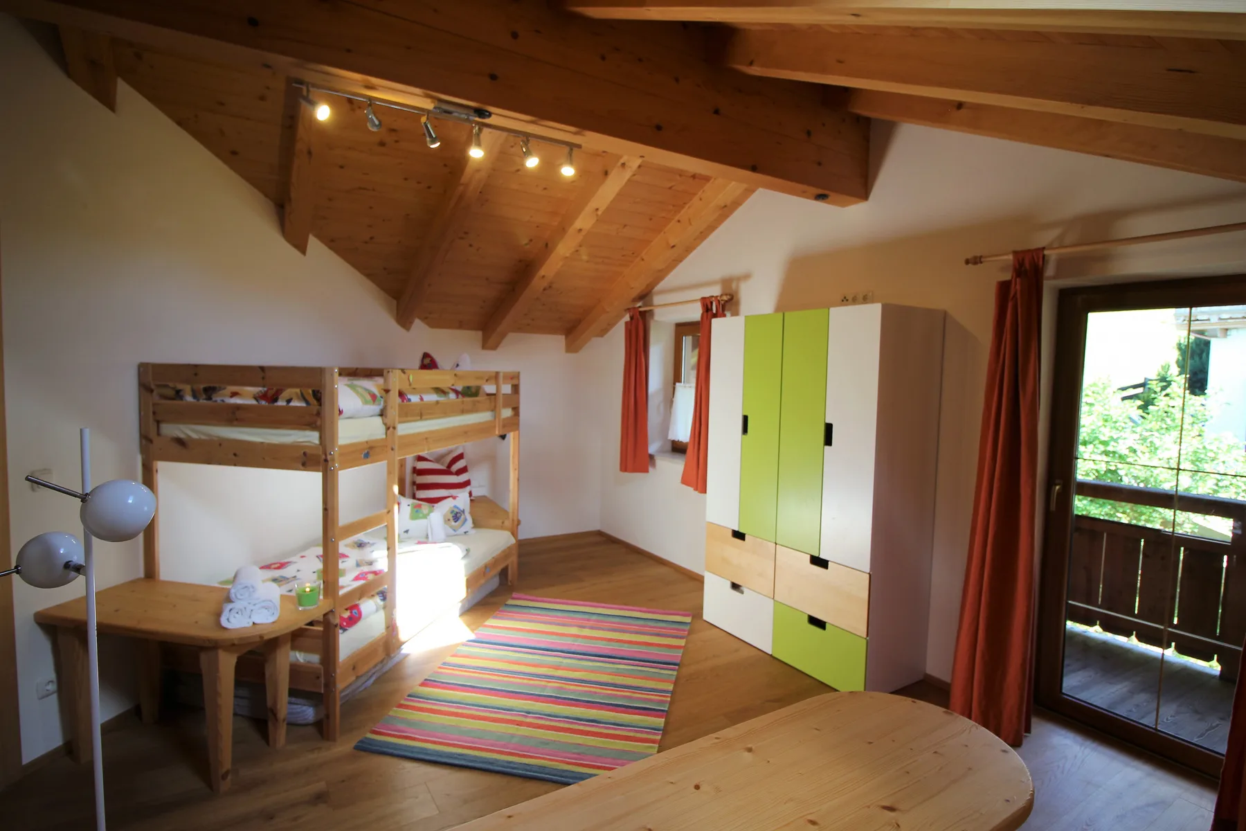 A bright bedroom with wooden bunk beds, a colorful rug, and a sloped timber ceiling.