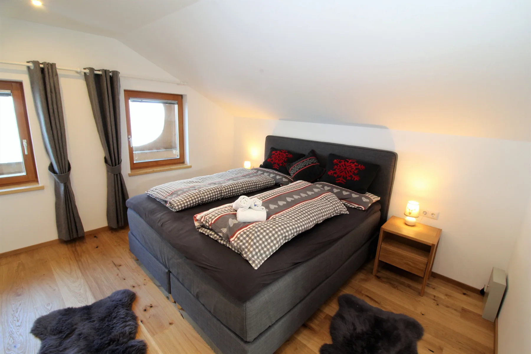 A bedroom with a bed dressed in dark bedding beside two curtained windows.