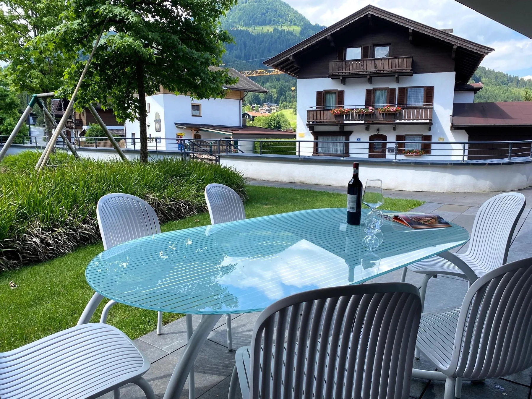Una zona de comedor al aire libre tiene una mesa de vidrio y asientos junto a un jardín.