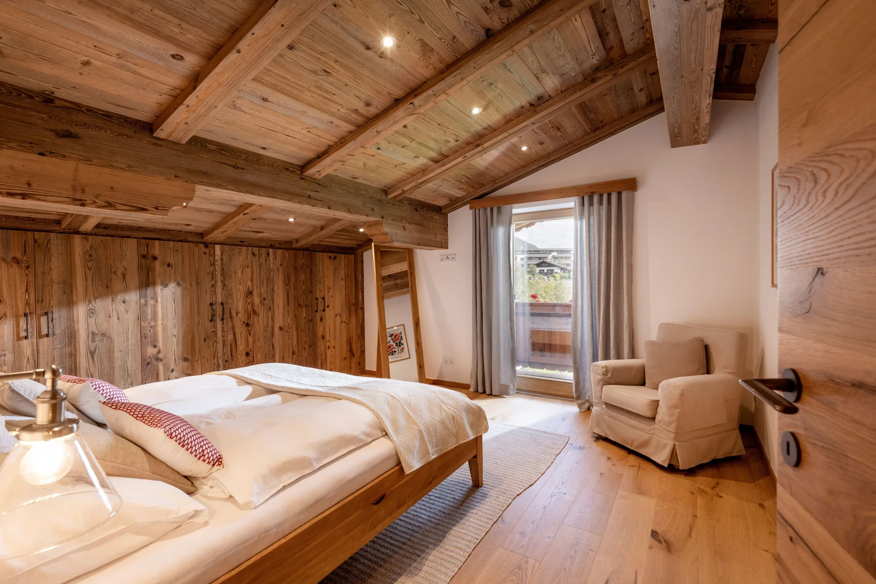 Un dormitorio con una cama de madera, techo inclinado de vigas y un sillón junto a la ventana.