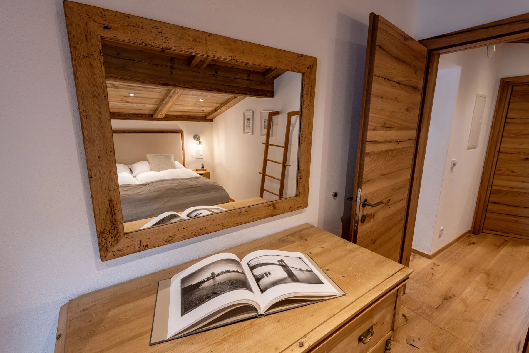 Una cómoda de madera con un libro abierto está bajo un espejo grande junto a la entrada de un dormitorio.