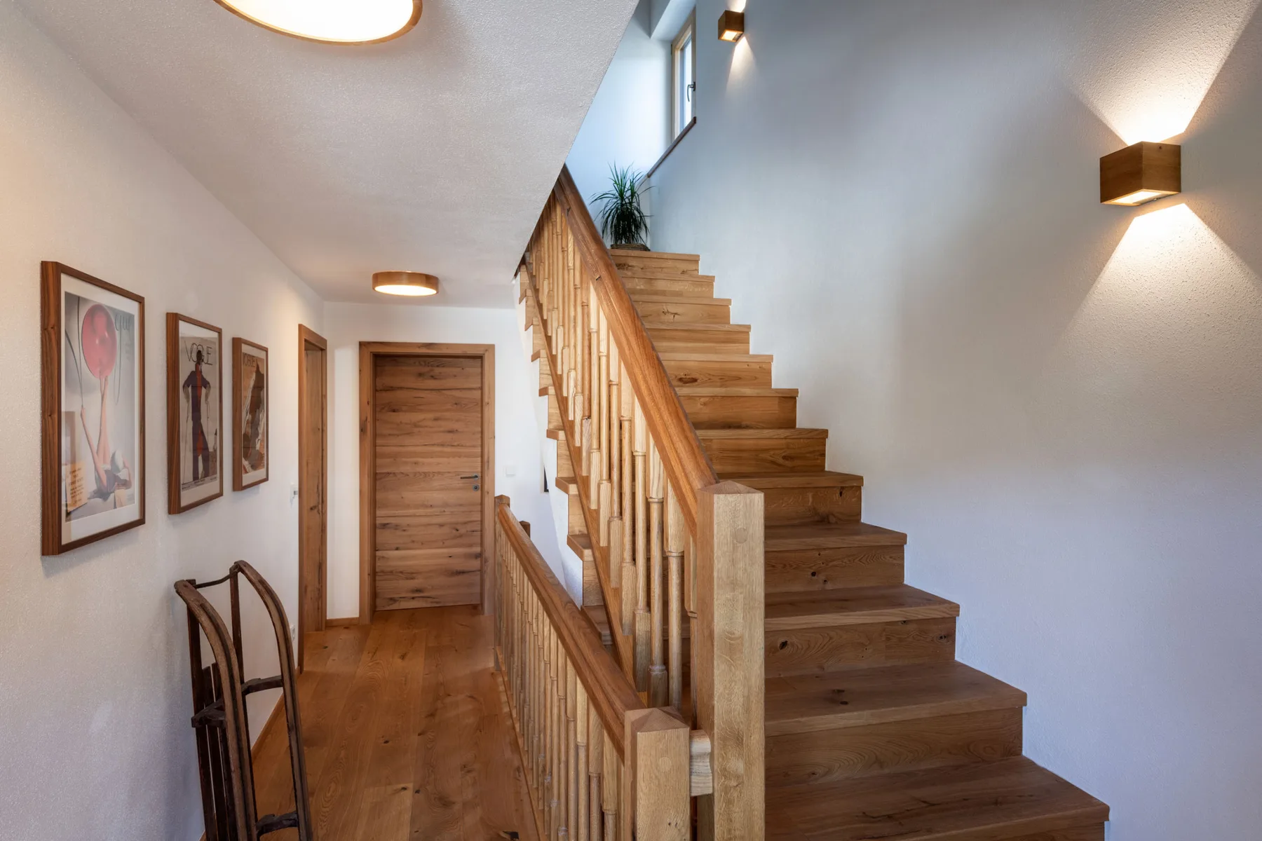 Una escalera de madera sube junto a un pasillo con cuadros enmarcados.