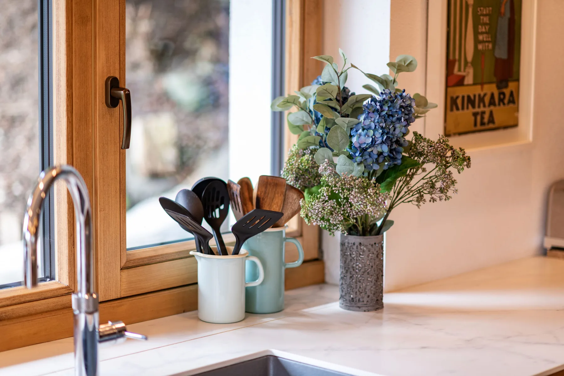 Una encimera de cocina junto a una ventana tiene utensilios y un jarrón con flores.
