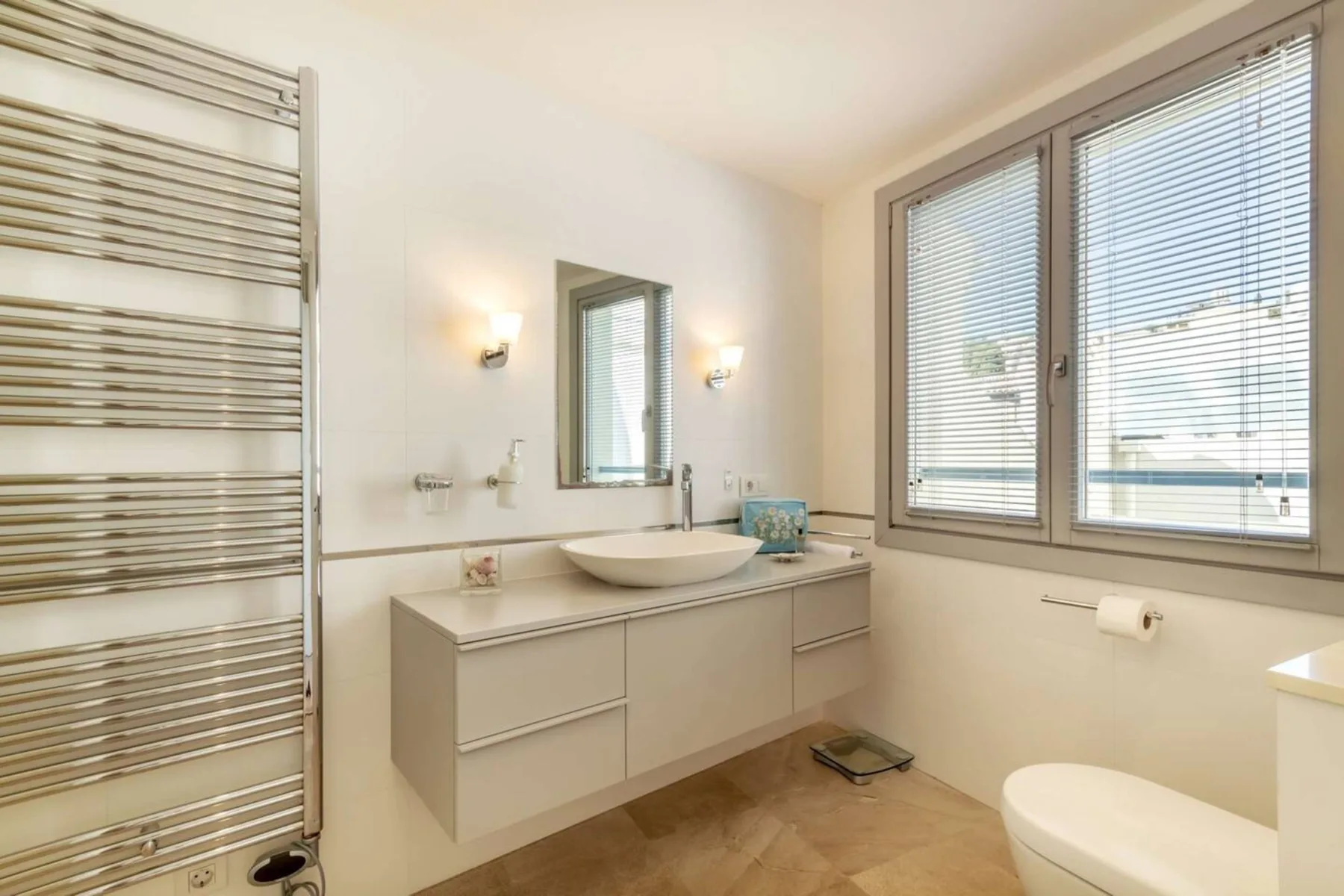 Un baño luminoso con tocador suspendido, lavabo sobre encimera y ventanas grandes.